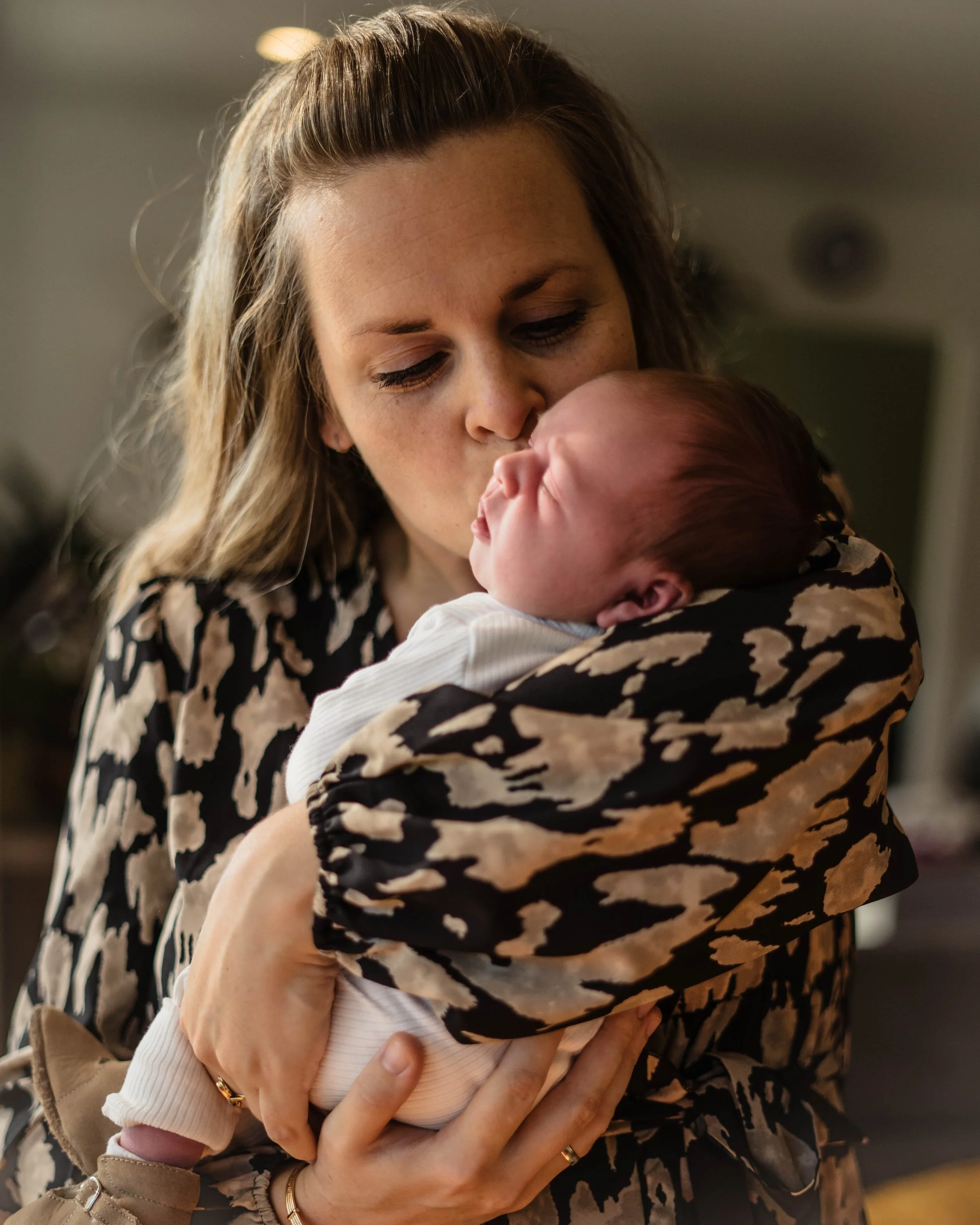 A woman with wavy light brown hair holding a sleeping baby, giving the baby a gentle kiss on the forehead in an indoor setting.