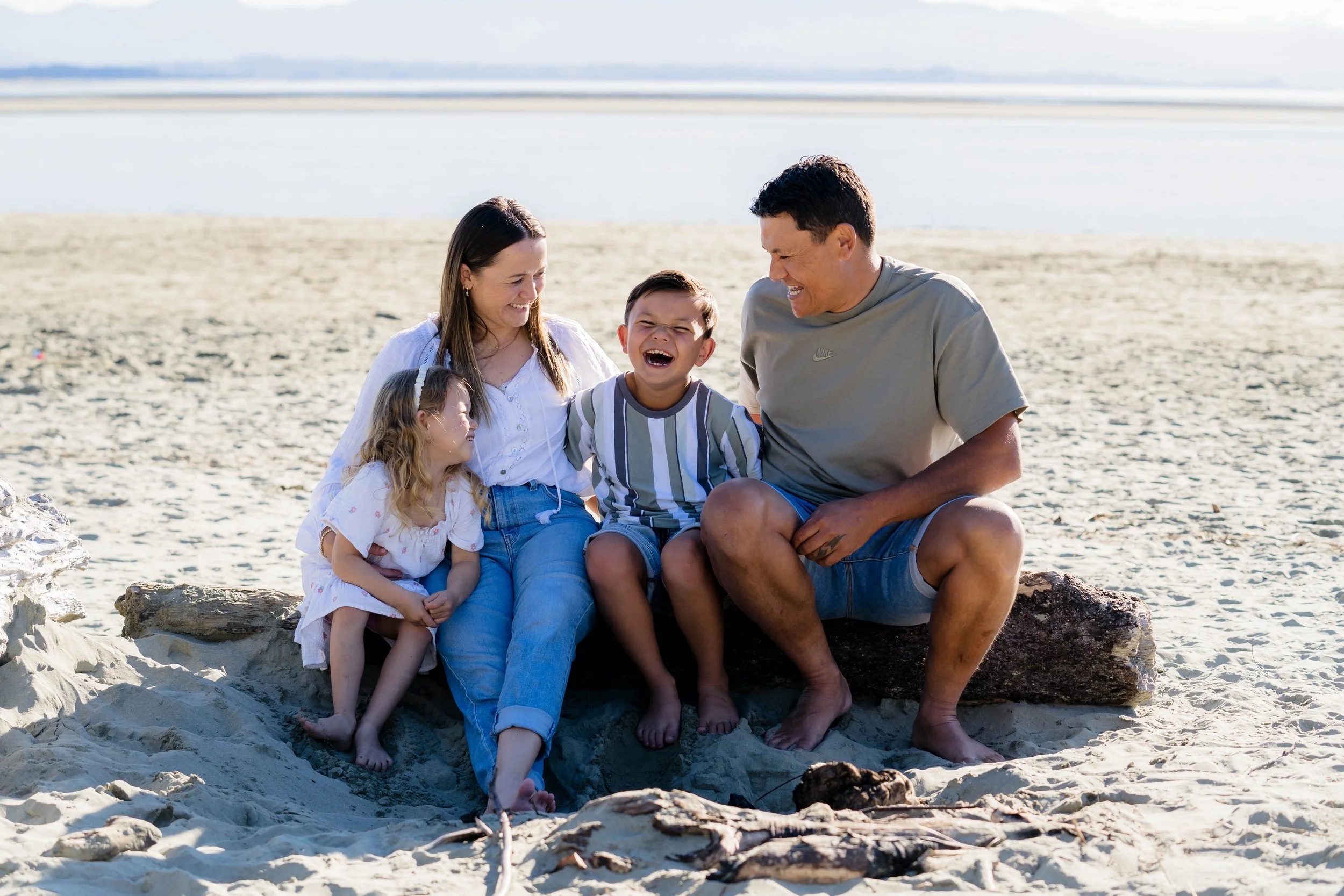 A happy family of five sitting on a log at the beach, laughing and enjoying their time together.