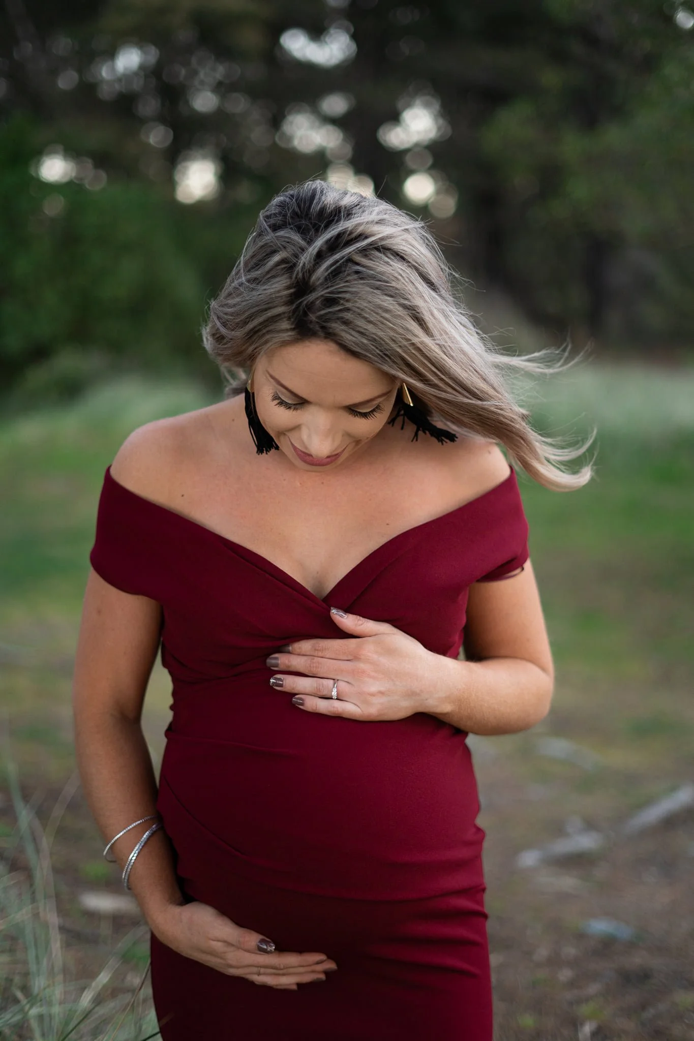 A woman in an off-the-shoulder burgundy dress standing outdoors, looking down with her hand on her stomach.