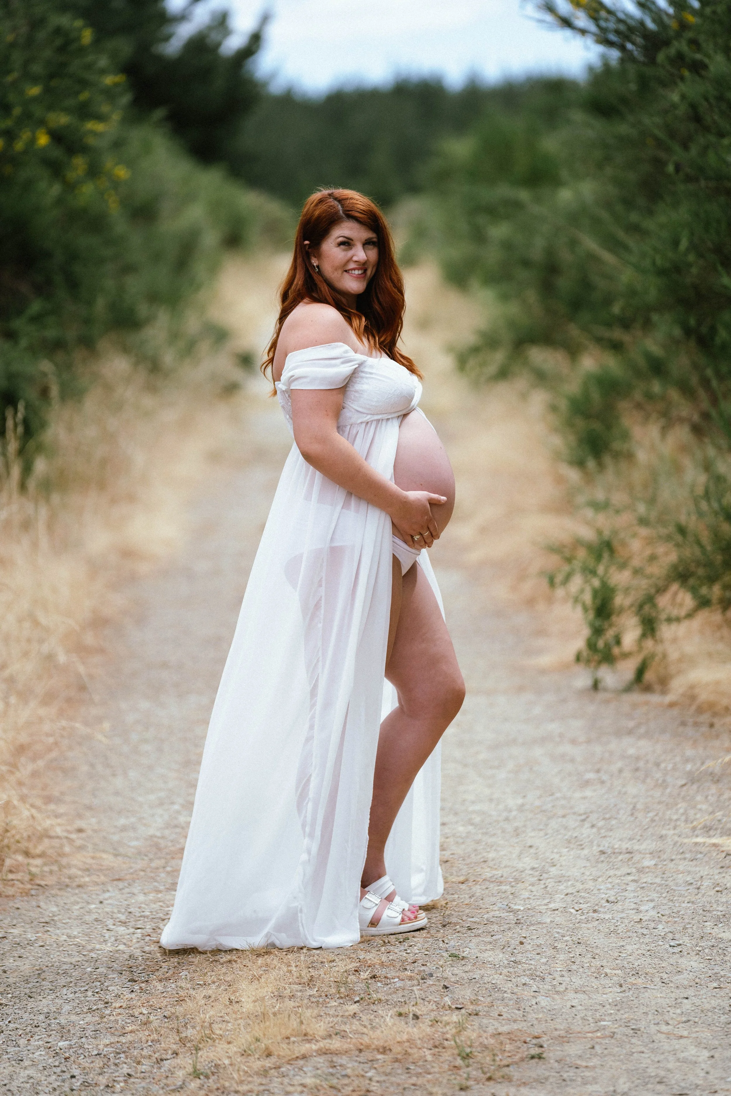 Pregnant woman in white dress standing on a dirt path surrounded by greenery, smiling and looking at the camera.