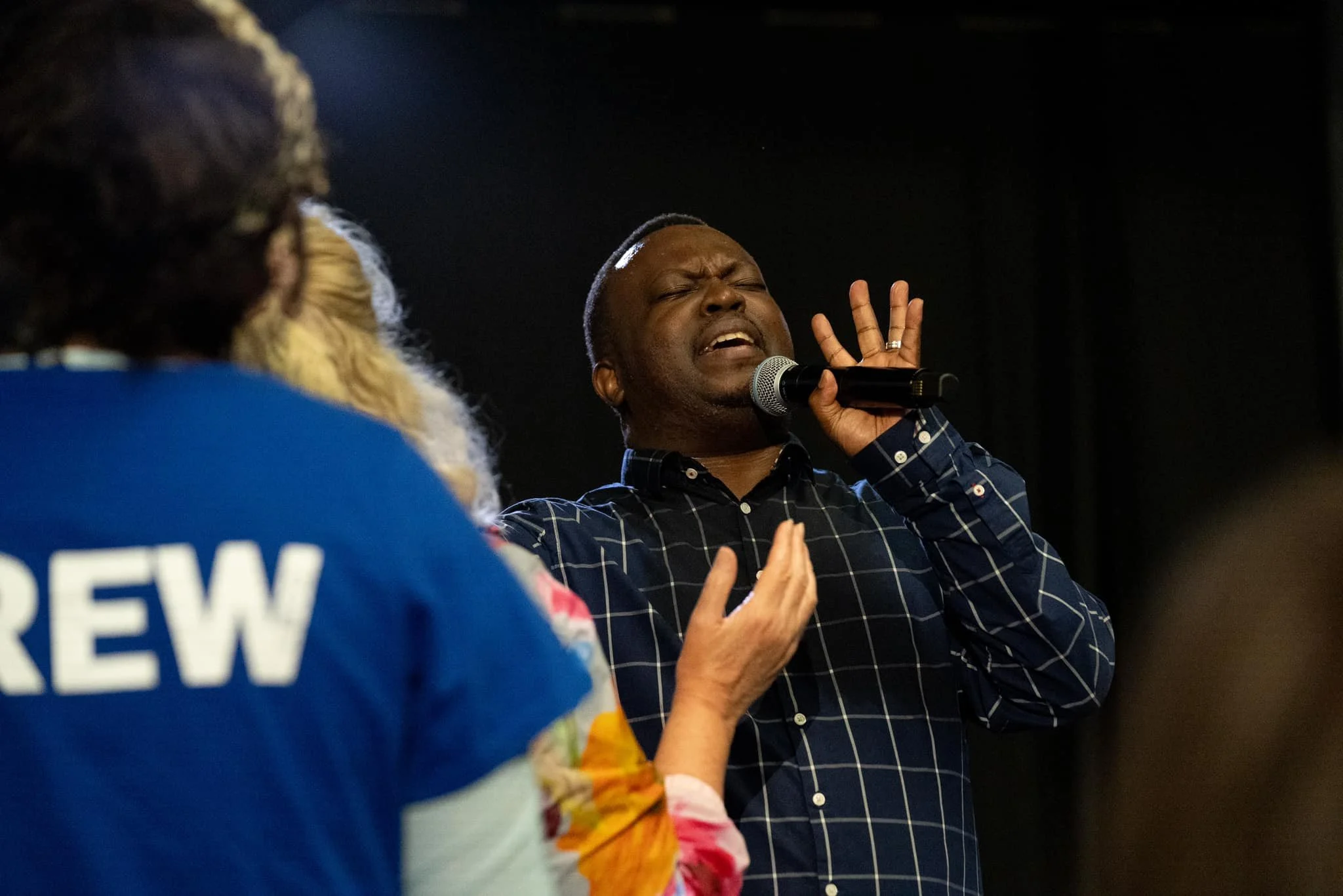 A man in a dark blue checkered shirt singing passionately with eyes closed, holding a microphone, while a woman in a blue shirt with the word 'CREW' on the back, gestures with her hand towards him.