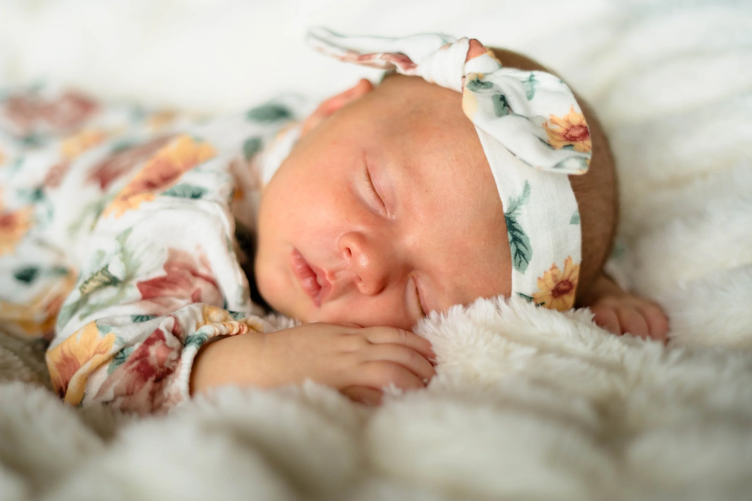 Close-up of a sleeping baby wearing a floral headband and matching outfit, resting on a soft, white blanket.