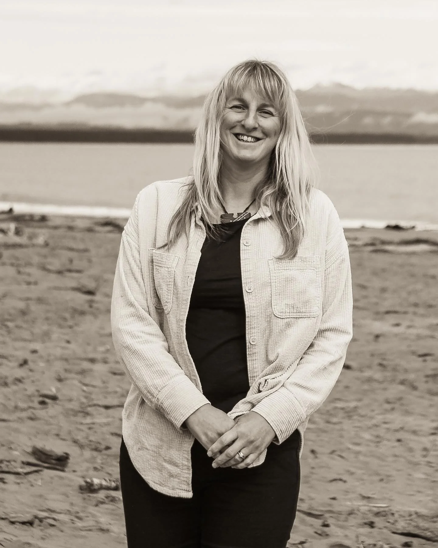 A woman smiling on a beach with a cloudy sky and water in the background.