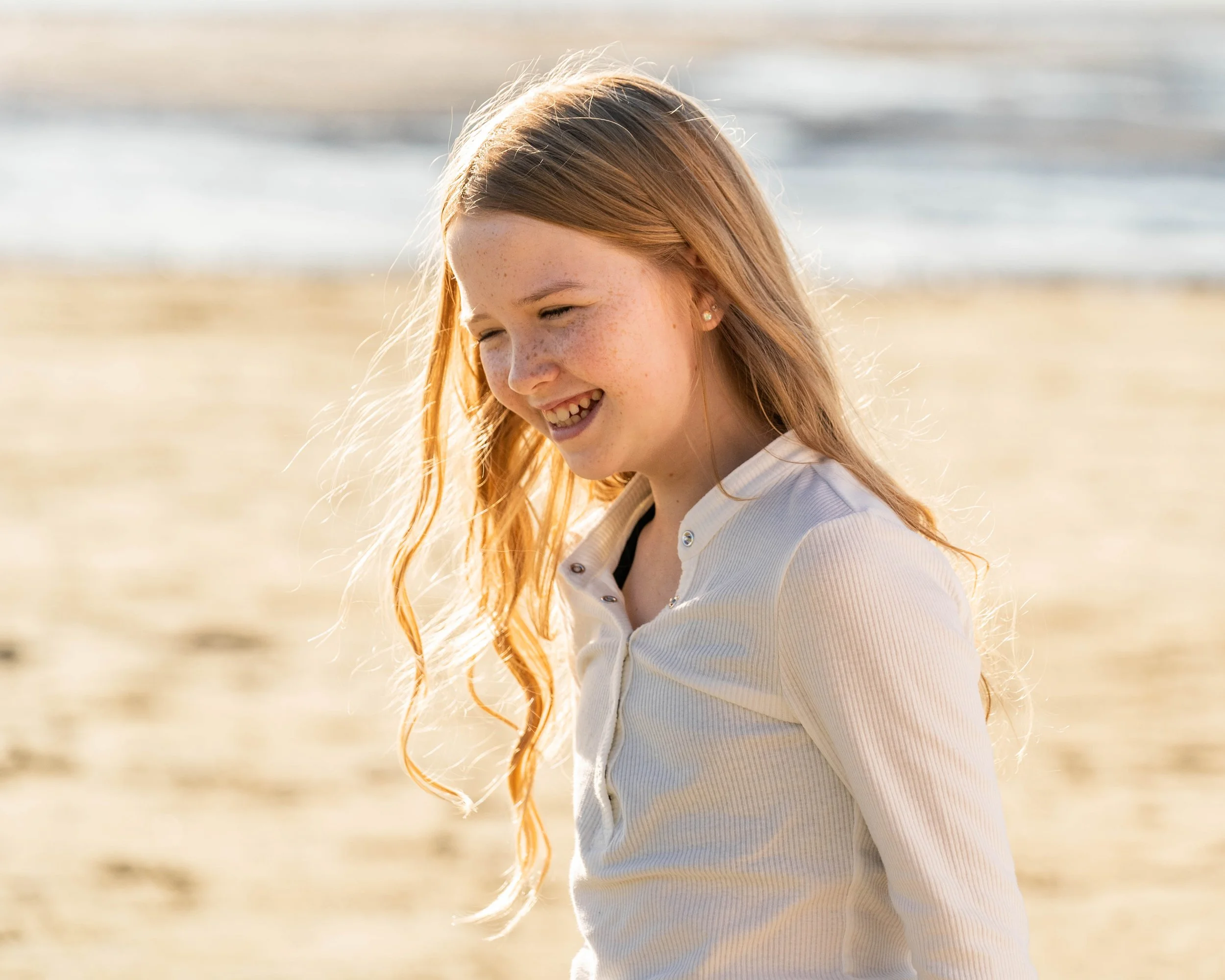 A young girl with red hair and freckles smiling and enjoying herself on the beach with ocean waves in the background.