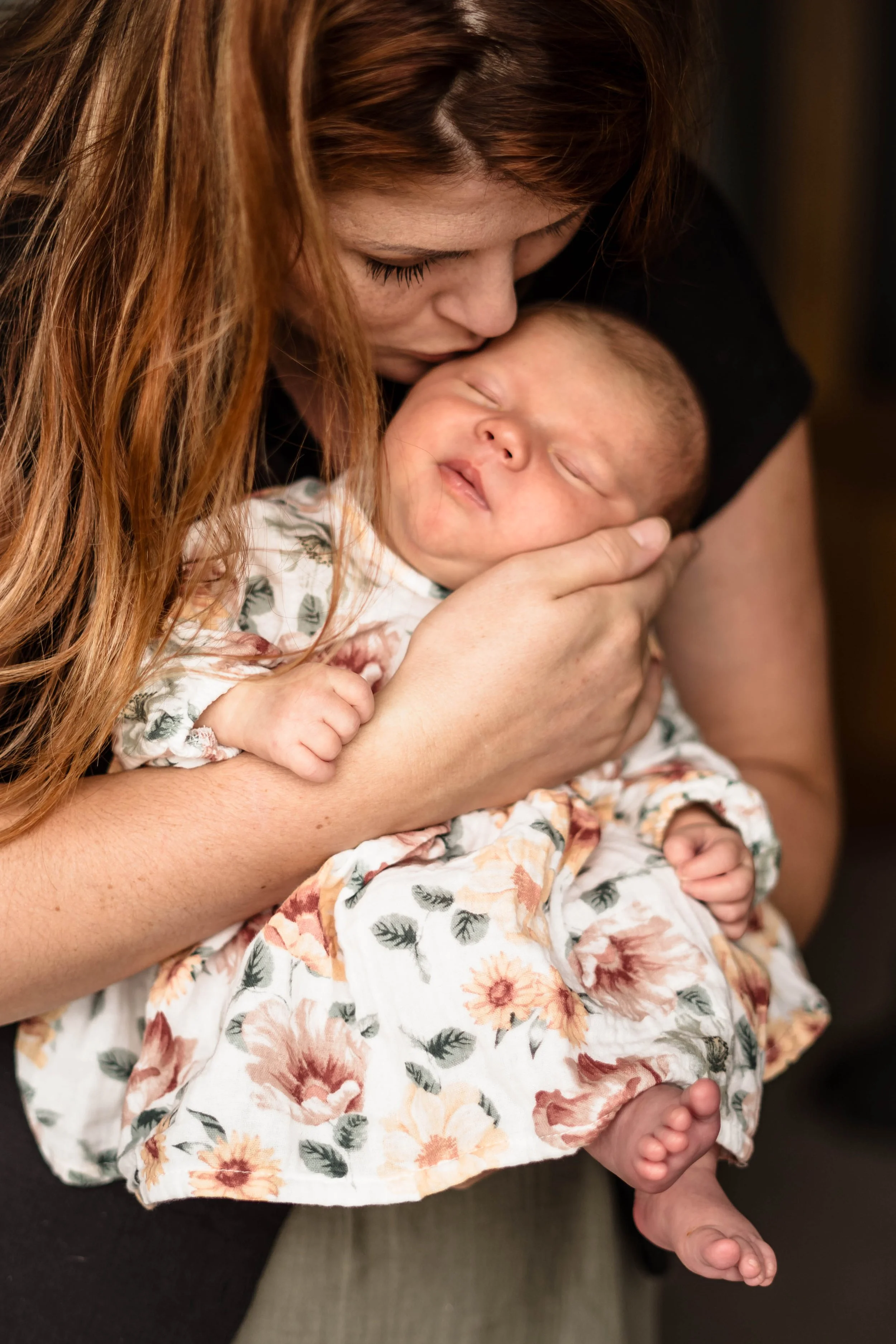 A woman with long red hair gently holds a sleeping baby, kissing the baby's forehead.