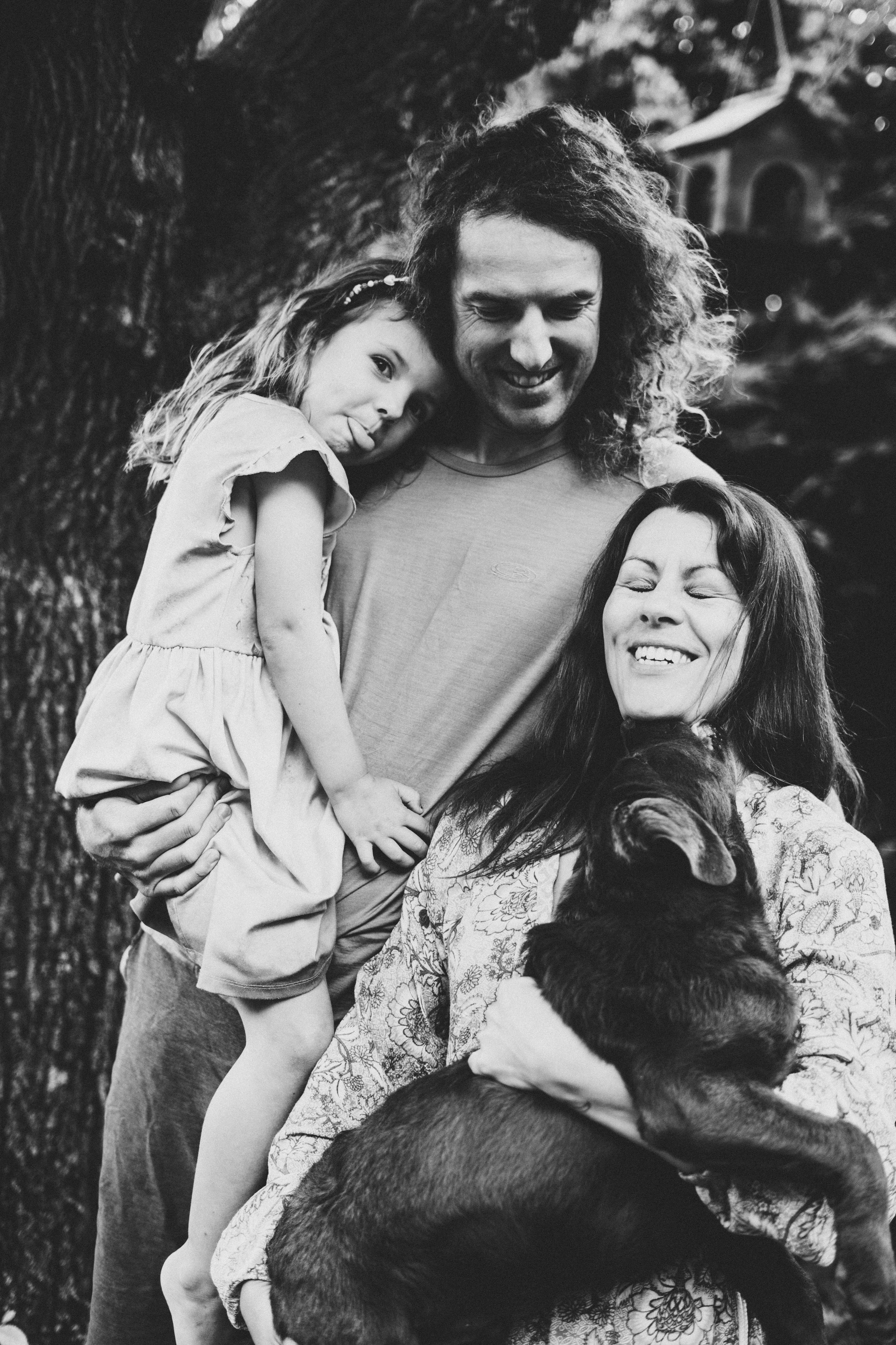 A family of three, with a young girl, a man, and a woman, smiling outdoors as the woman holds a big puppy. The girl is leaning her head on the man's shoulder, and the family is near a tree.