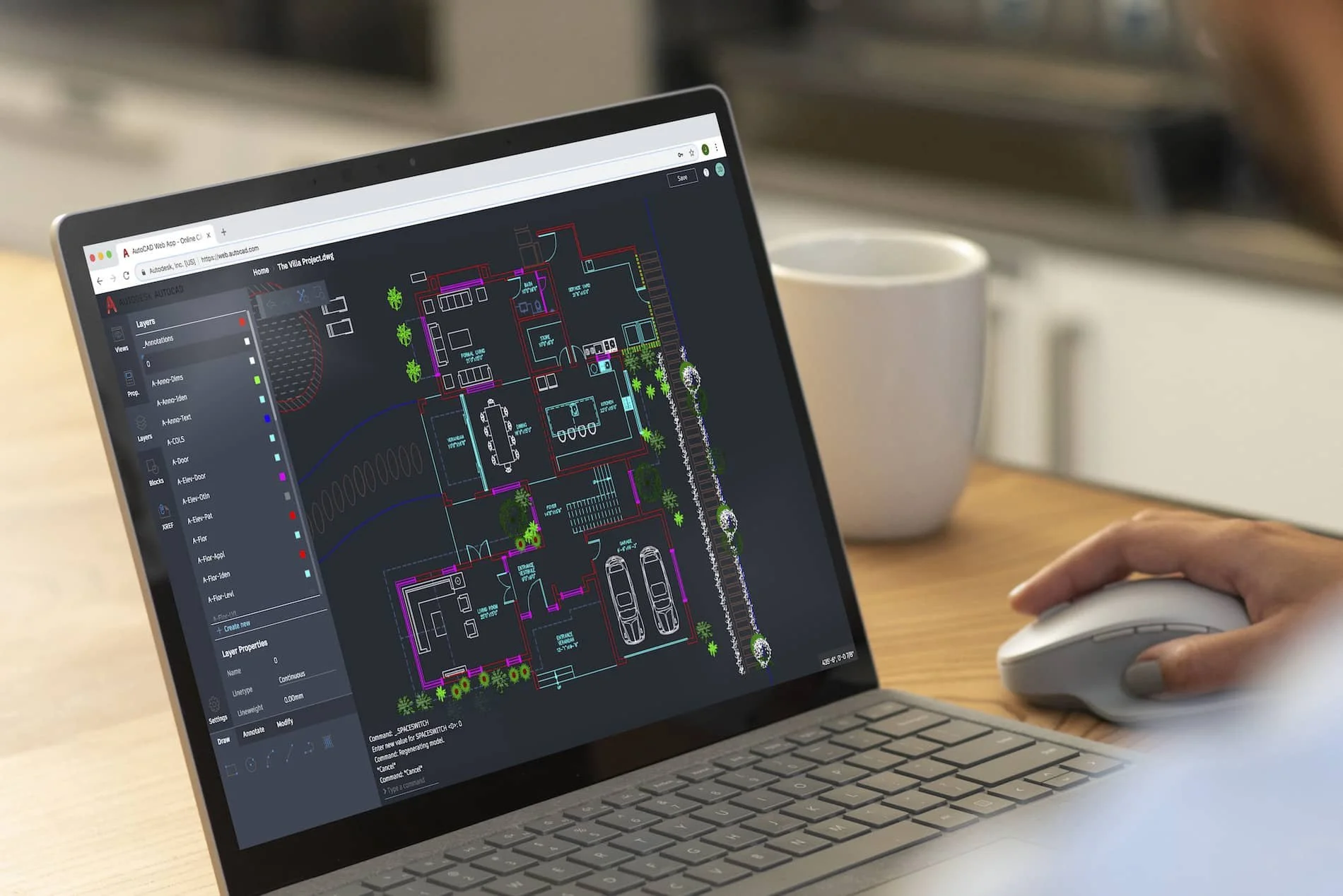A person using a laptop with a CAD architectural drawing of a house displayed on the screen, sitting at a wooden desk with a white mug nearby.