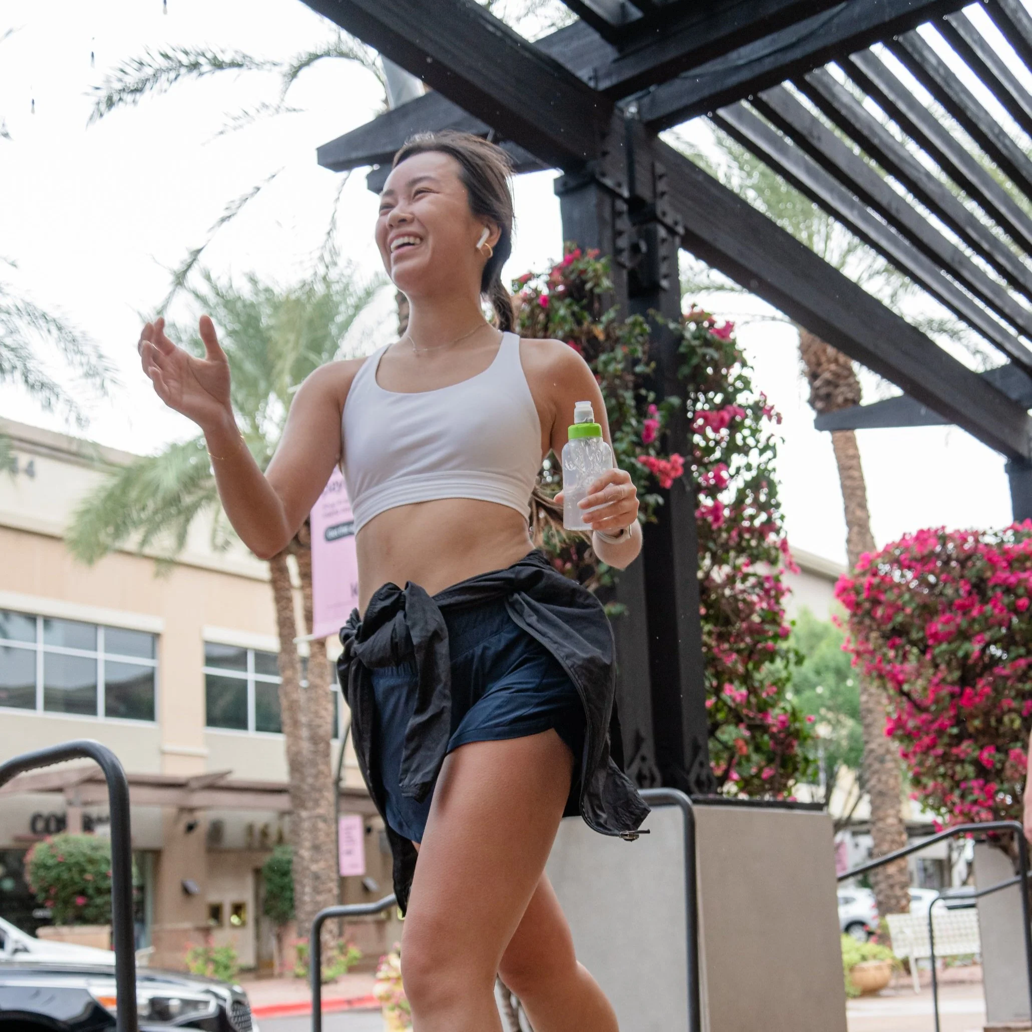 A woman jogging outdoors, smiling, holding a water bottle, wearing athletic clothing, with colorful flowering plants and palm trees in the background.