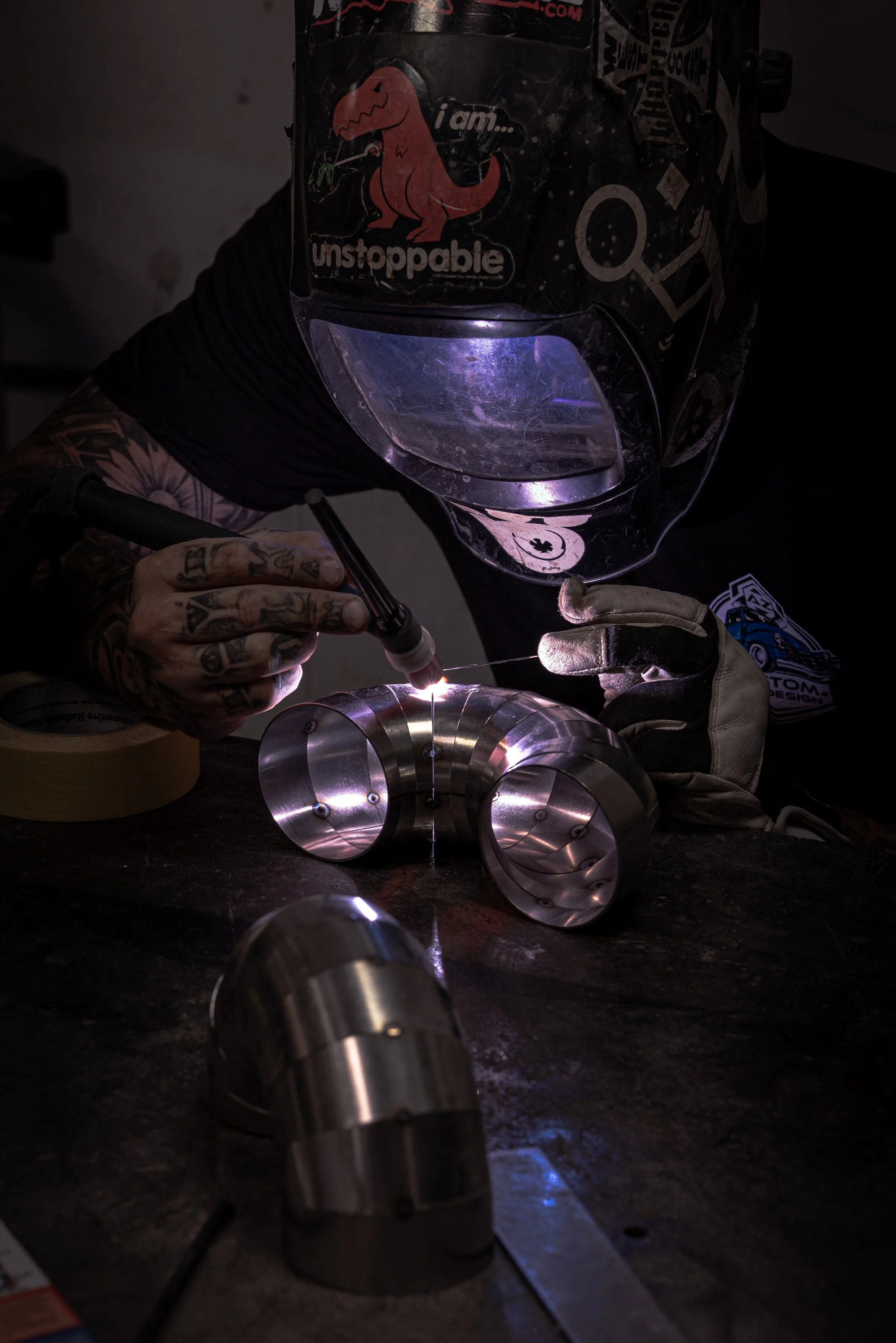 A person wearing a welding helmet and gloves welding a metallic pipe assembly in a workshop. The worker's hands are guiding a welding tool, with sparks and bright light emitted during the welding process.