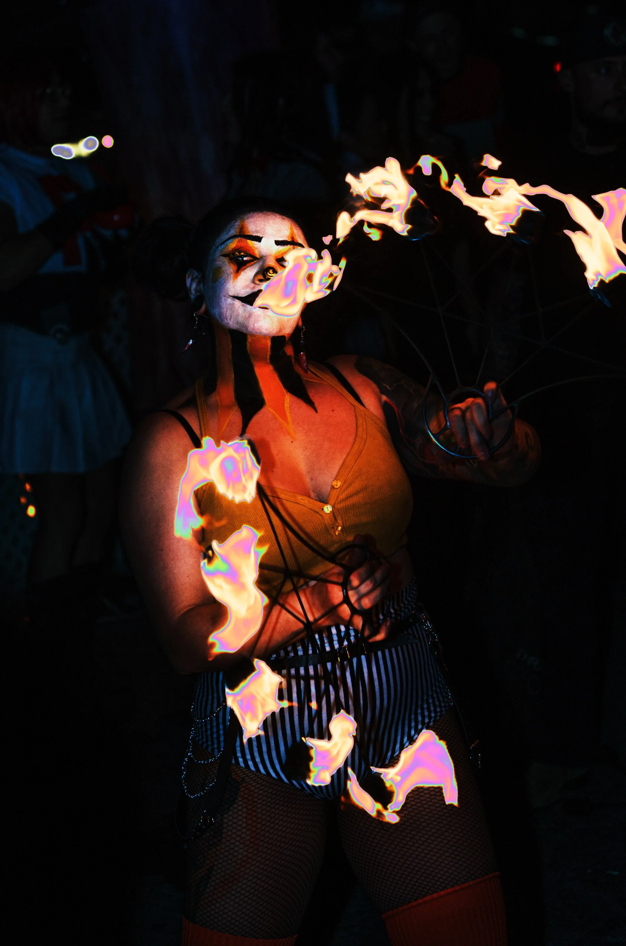 Person dressed as a clown with painted face, burning fire poi, and colorful costume performing in a dark setting.