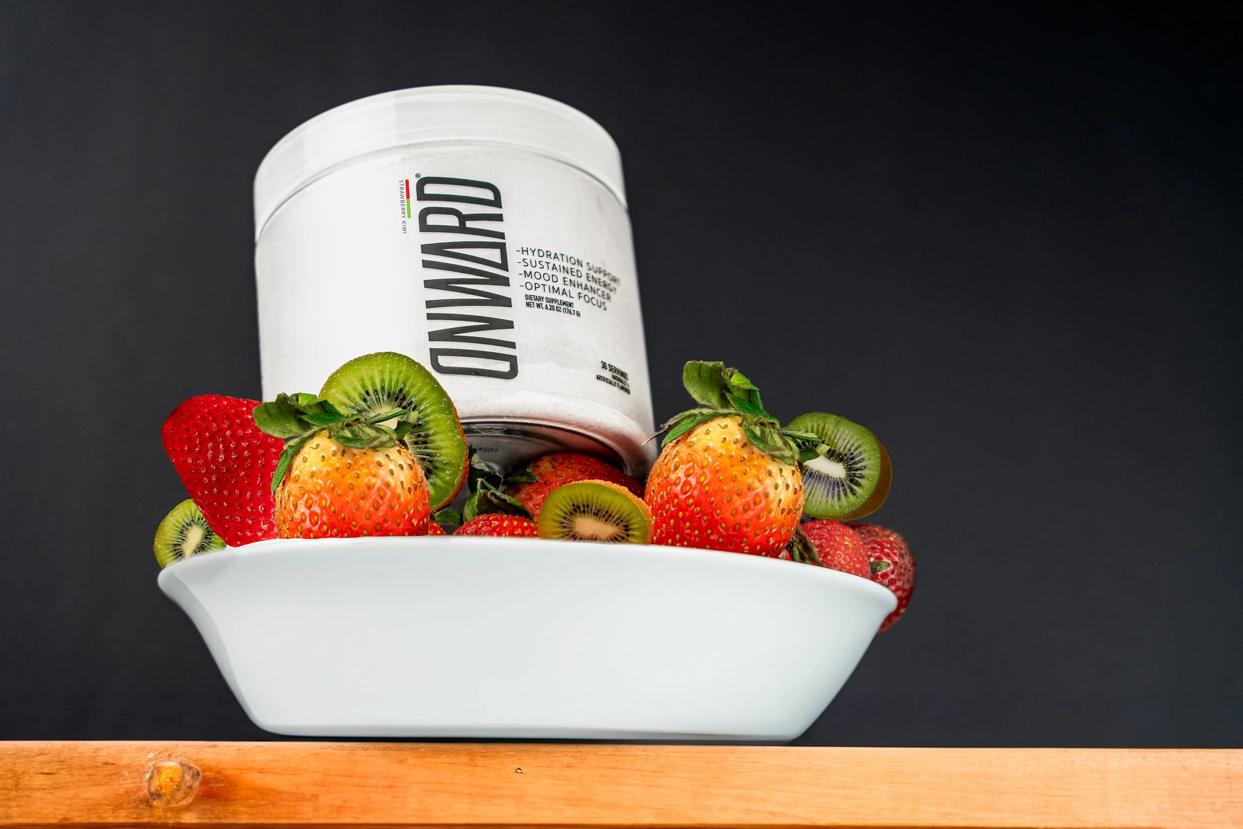 White dish filled with strawberries and sliced kiwis, with a container labeled 'DOWN MORE' balanced on top of the fruits, against a black background.