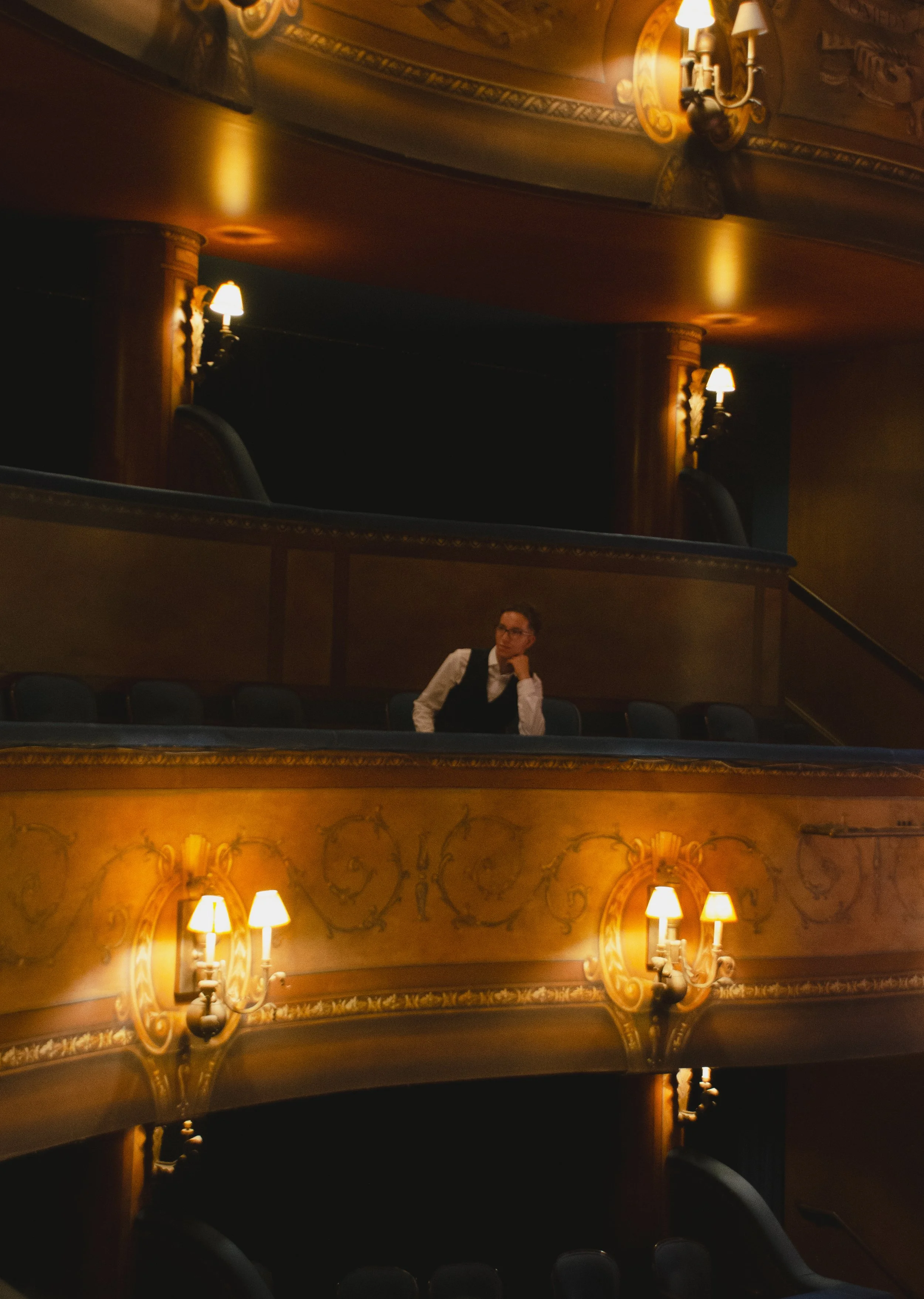 A person in formal attire sitting alone in a vintage theater balcony, with ornate wooden and gold decor, and warm yellow lighting from wall sconces.