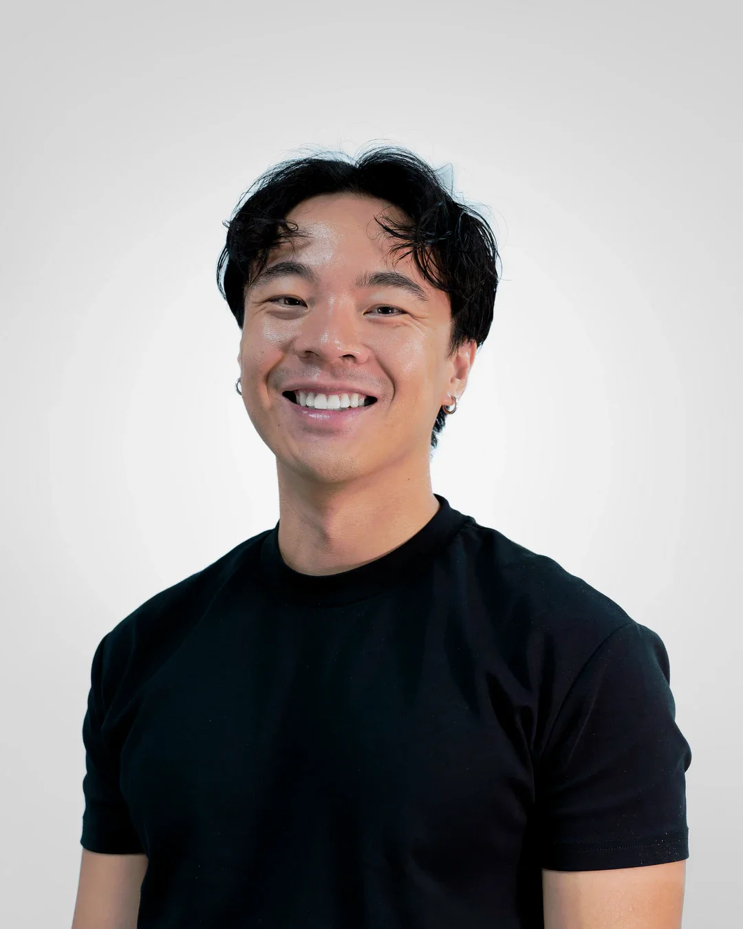 A smiling young man with black hair, wearing a black t-shirt, standing against a plain white background.