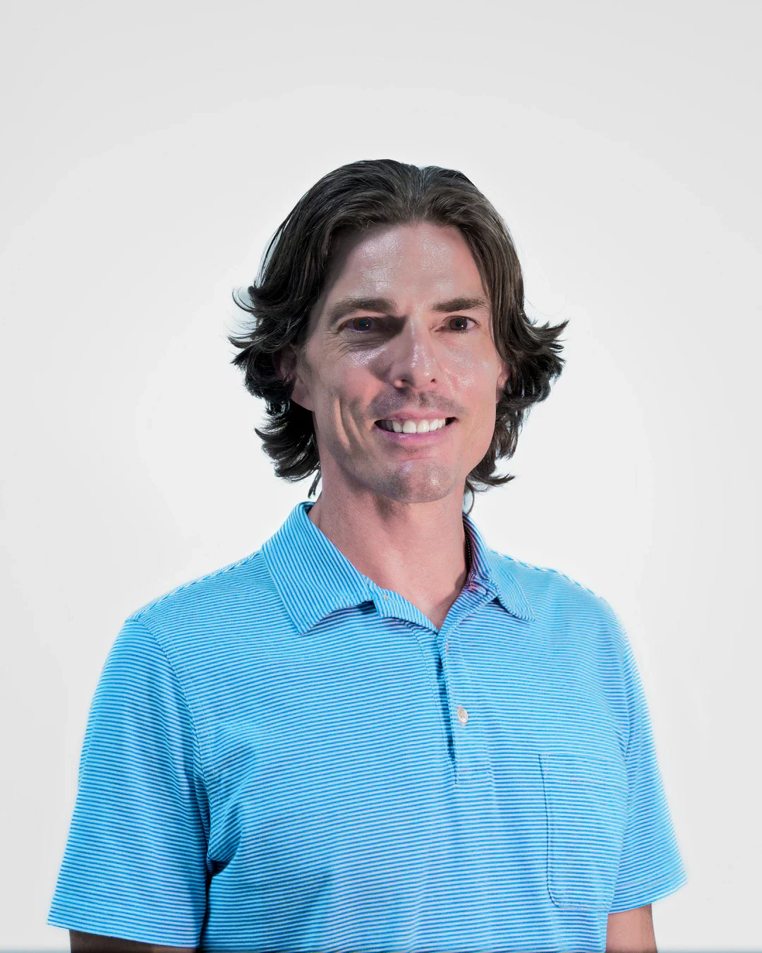 A middle-aged man with long, dark hair, wearing a light blue, striped polo shirt, smiling against a plain white background.
