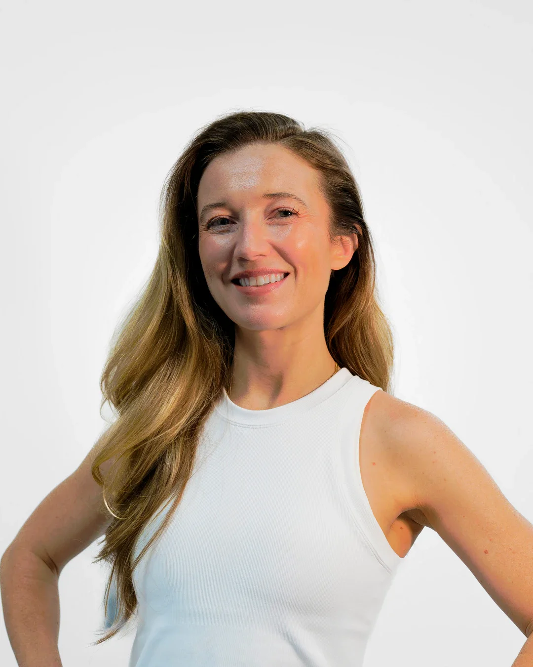 Woman with long, wavy hair wearing a white sleeveless top, smiling and standing against a plain white background.