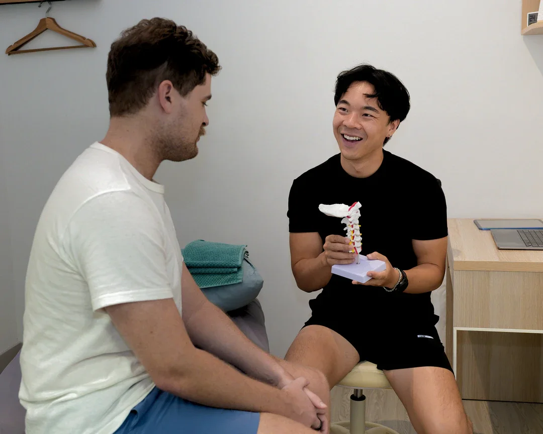 A man with dark hair and Asian features is smiling and holding a model of a human spine. He is sitting in a medical office with a neutral-colored wall and a small wooden desk nearby. A second man with light hair and skin is sitting across from him, listening attentively.