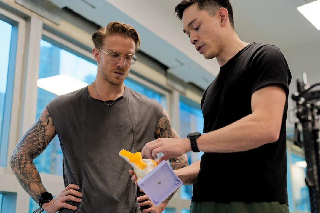 Two men are in a gym, one with tattoos on his arms wearing glasses and a sleeveless shirt, and the other in a black T-shirt, examining a foam roller or massage device.