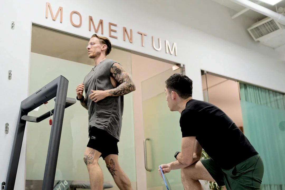 A man with tattoos running on a treadmill in a gym while another man in black shorts and a black t-shirt crouches nearby, observing or coaching. The gym has a sign that reads 'MOMENTUM' on the wall.