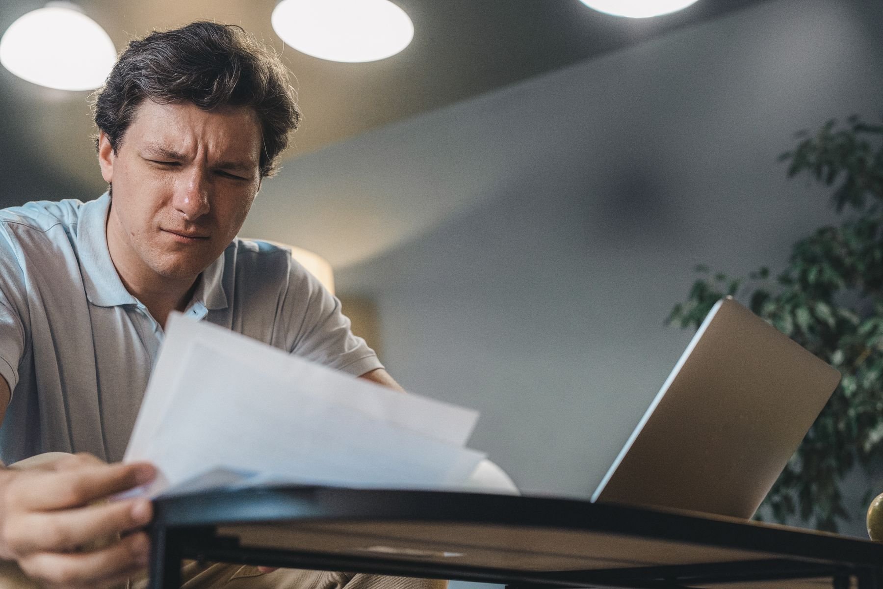 A man reading printed documents beside a laptop.