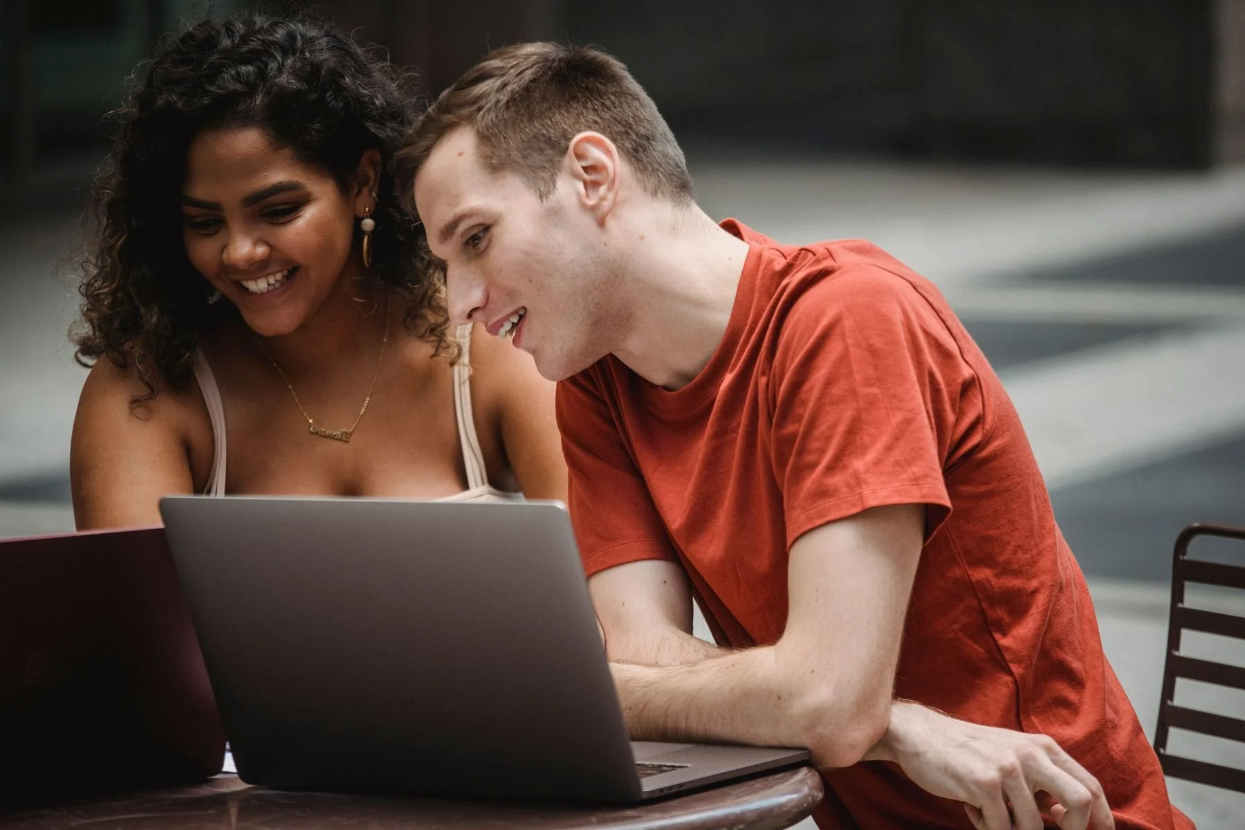 A smiling couple leaning toward a laptop at an outdoor café.