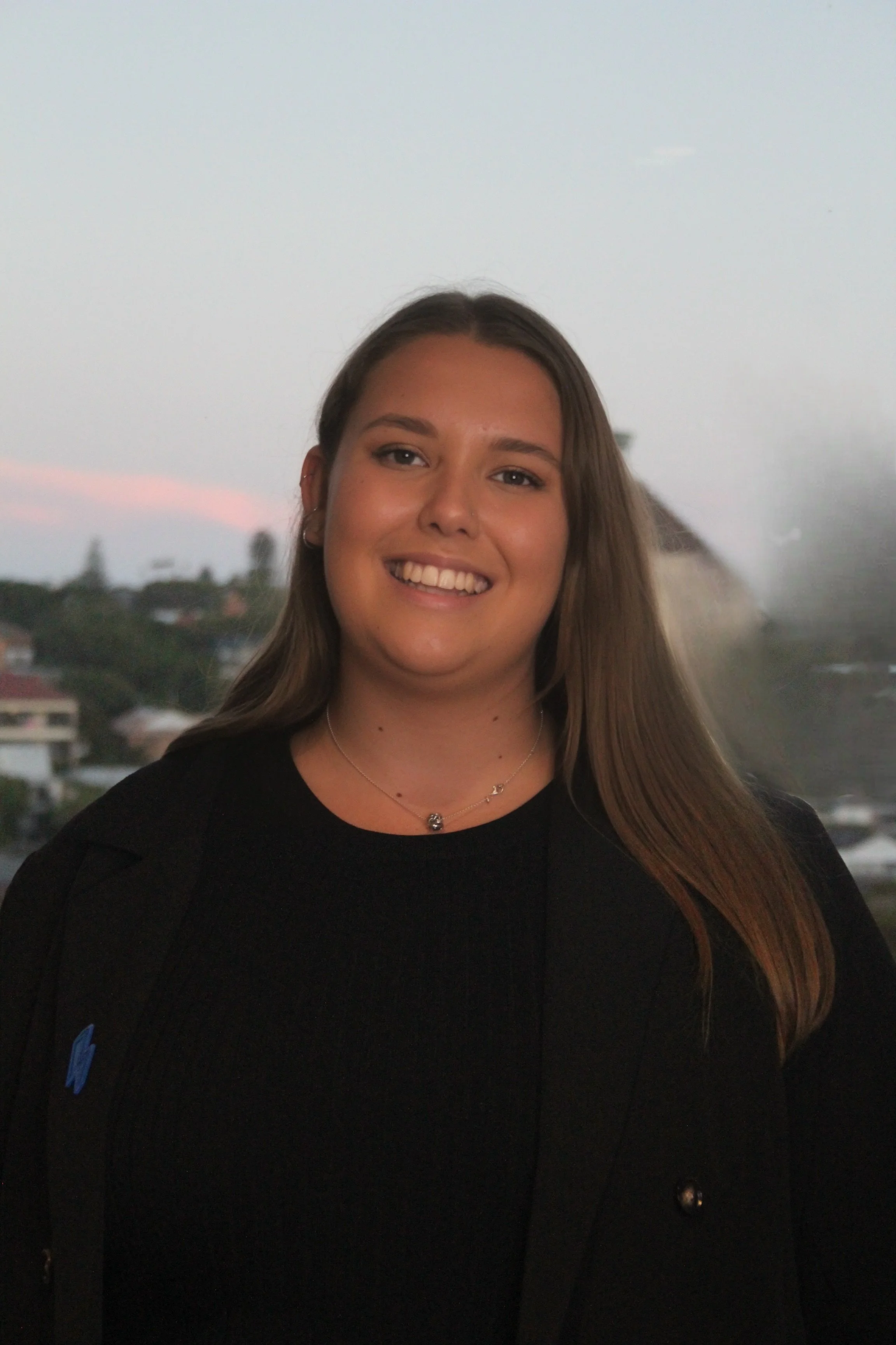 A smiling woman with long hair wearing a black top and a necklace, standing outdoors with blurred trees in the background.
