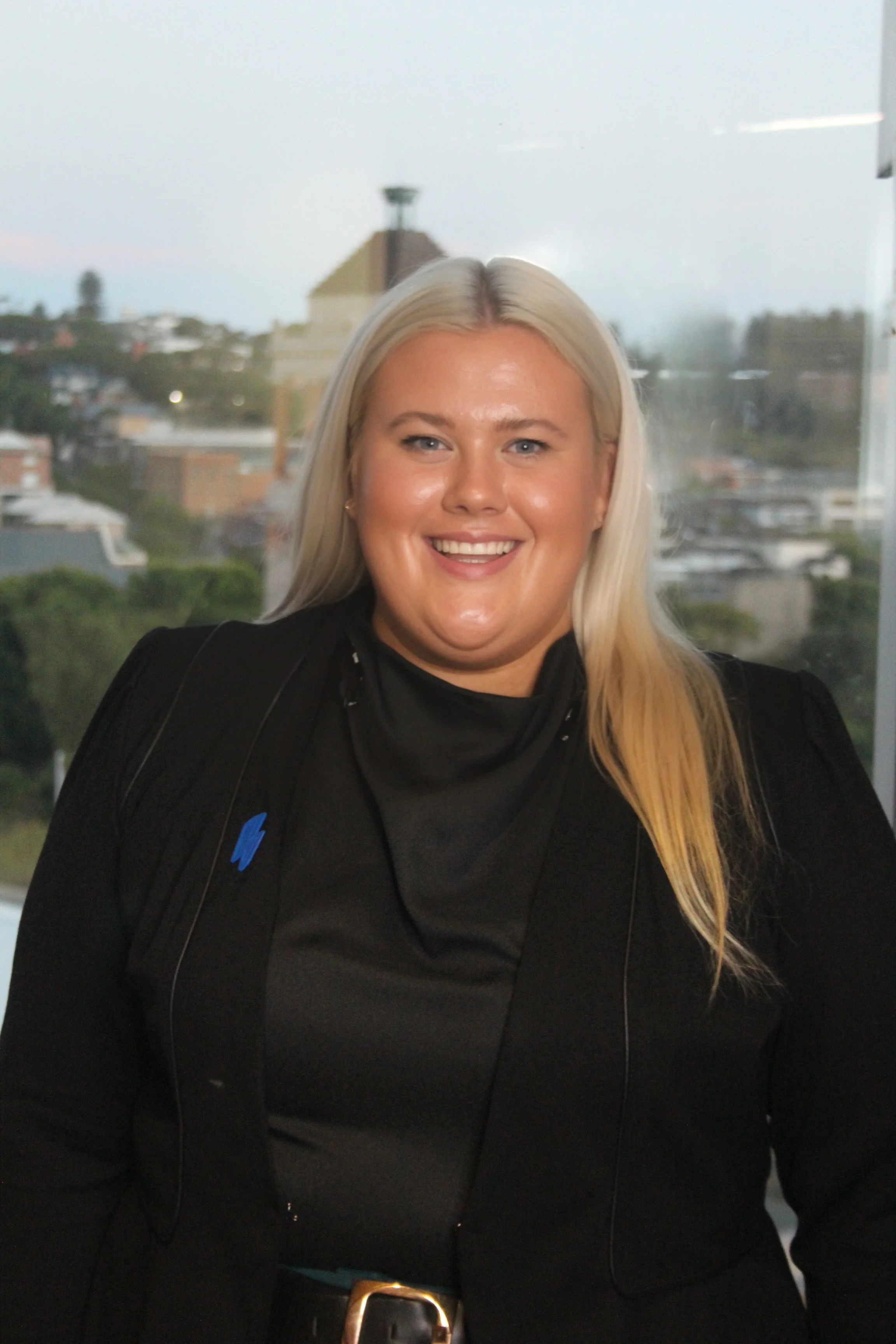 Person smiling indoors with cityscape in the background; wearing a black outfit and long blonde hair.
