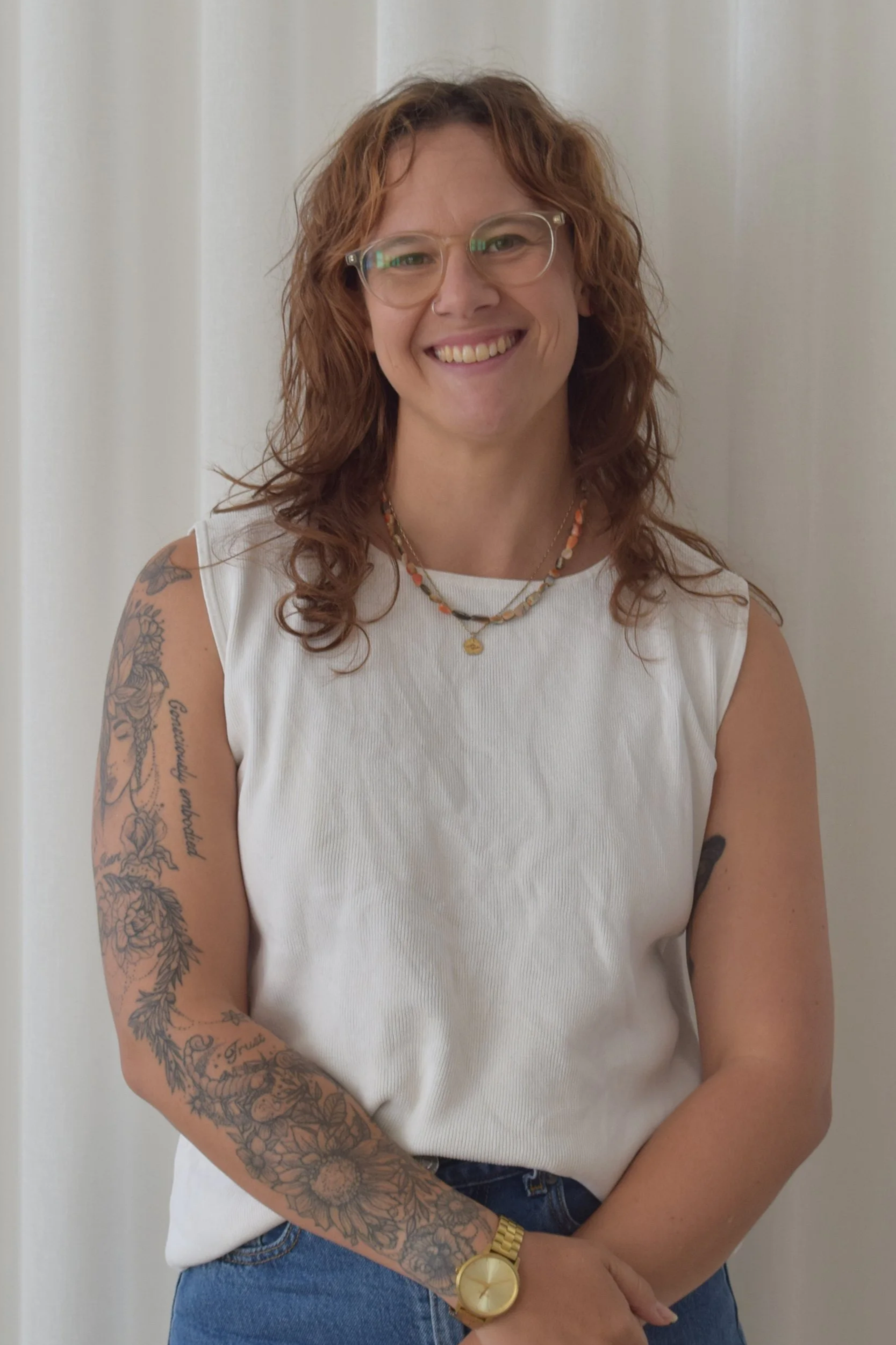 Person wearing a white top with a tattooed arm. They have brown hair and wear glasses.