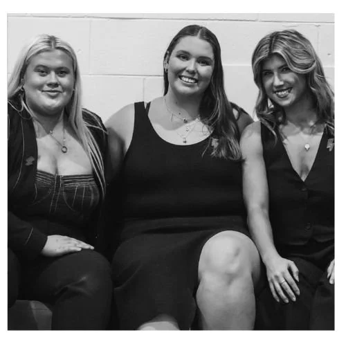 Three women sitting together, smiling, in a black and white photo. They are wearing formal attire and have long hair. The background is a plain wall.