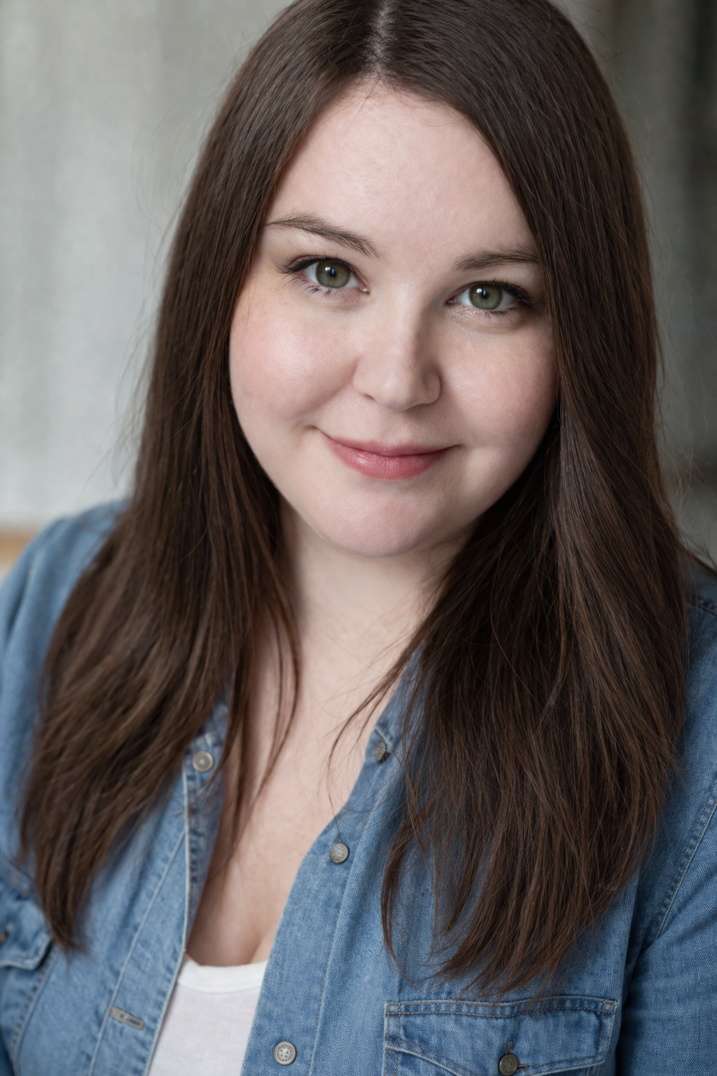 A young woman with long brown hair, green eyes, and a gentle smile, wearing a denim jacket and white top, looking at the camera.