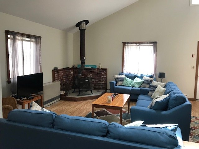 Living room with blue sofas, a brick fireplace with a black wood stove, a wooden coffee table, a small TV on a stand, and windows with curtains.