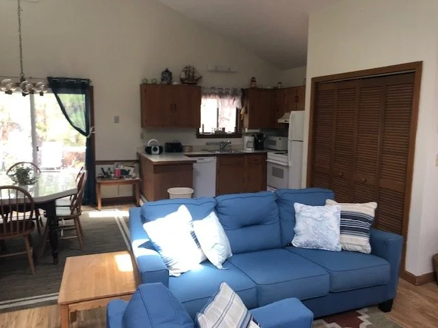Living room with blue sofa and pillows, adjacent dining area with wooden table and chairs, kitchen with wooden cabinets, white appliances, and curtains over window; sunlight streaming in.