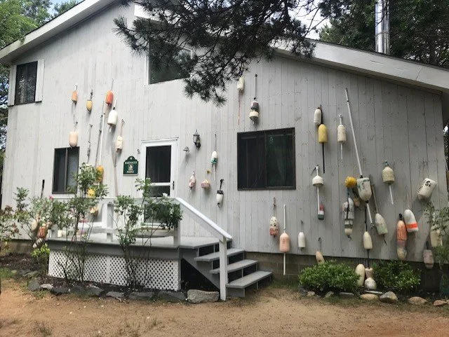 A white house with buoys hanging on the exterior wall and around the porch area, with a couple of windows and a small set of stairs leading to the door.
