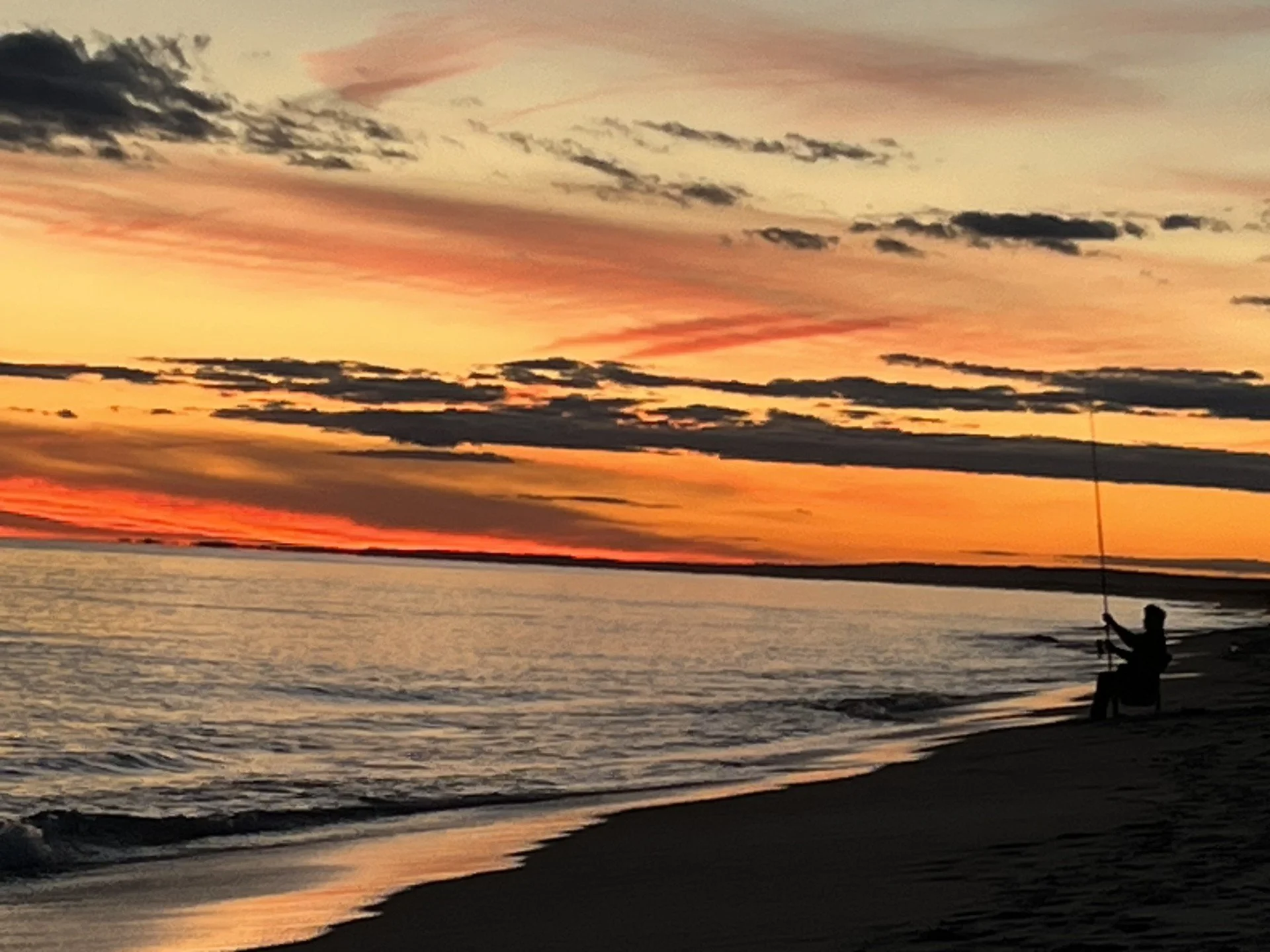 Silhouette of a person fishing on a beach at sunset with colorful sky and calm water.
