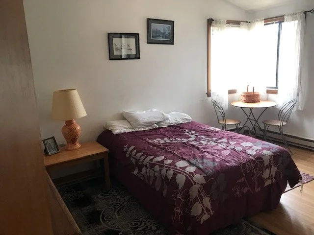 A bedroom with a bed covered in a purple floral quilt, two pillows, a wooden nightstand with a lamp, and framed pictures on the wall. There's a window with curtains and a small table with two chairs nearby.