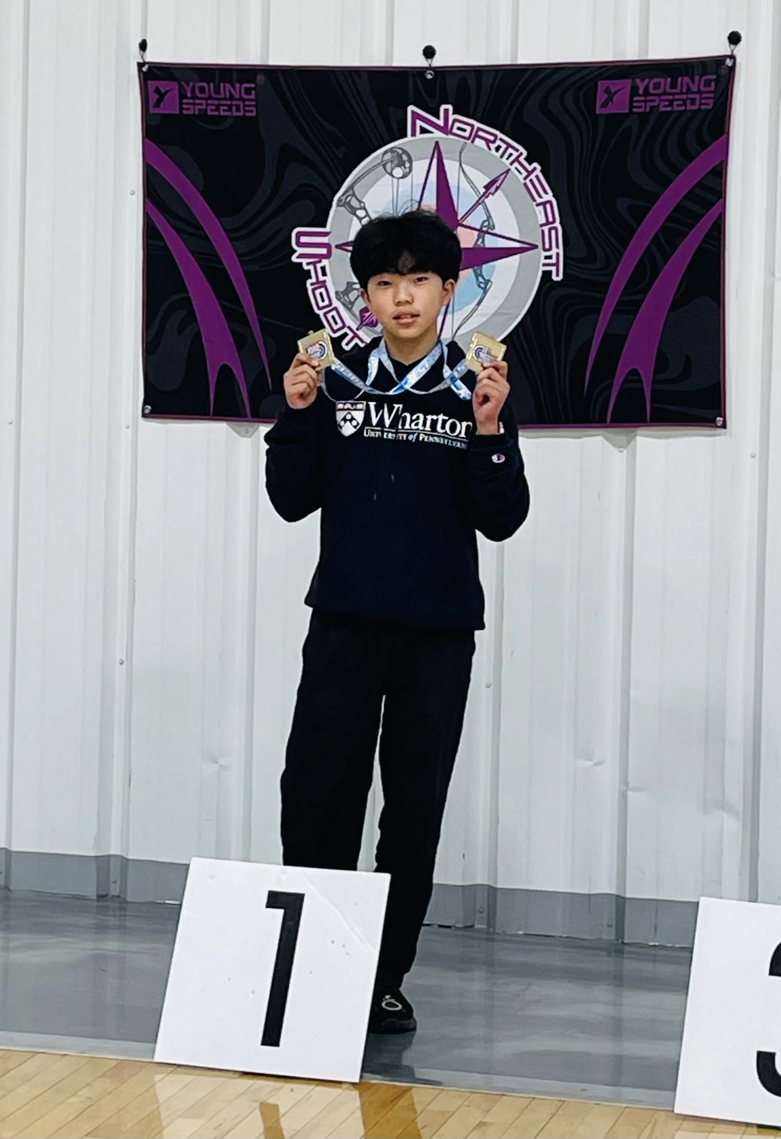 A young boy standing on a winner's podium holding two medals, in front of a banner with a compass graphic, the words "North East Up-Down" and "Young Speed".