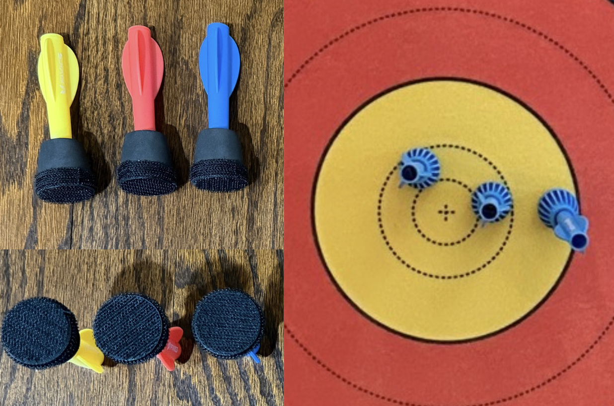 Three darts with different colored handles (yellow, pink, blue) and foam grips are shown from top and bottom views. The darts are placed on a wooden surface, with three matching foam targets in front of them. There are also three darts stuck to a yellow and red target paper.
