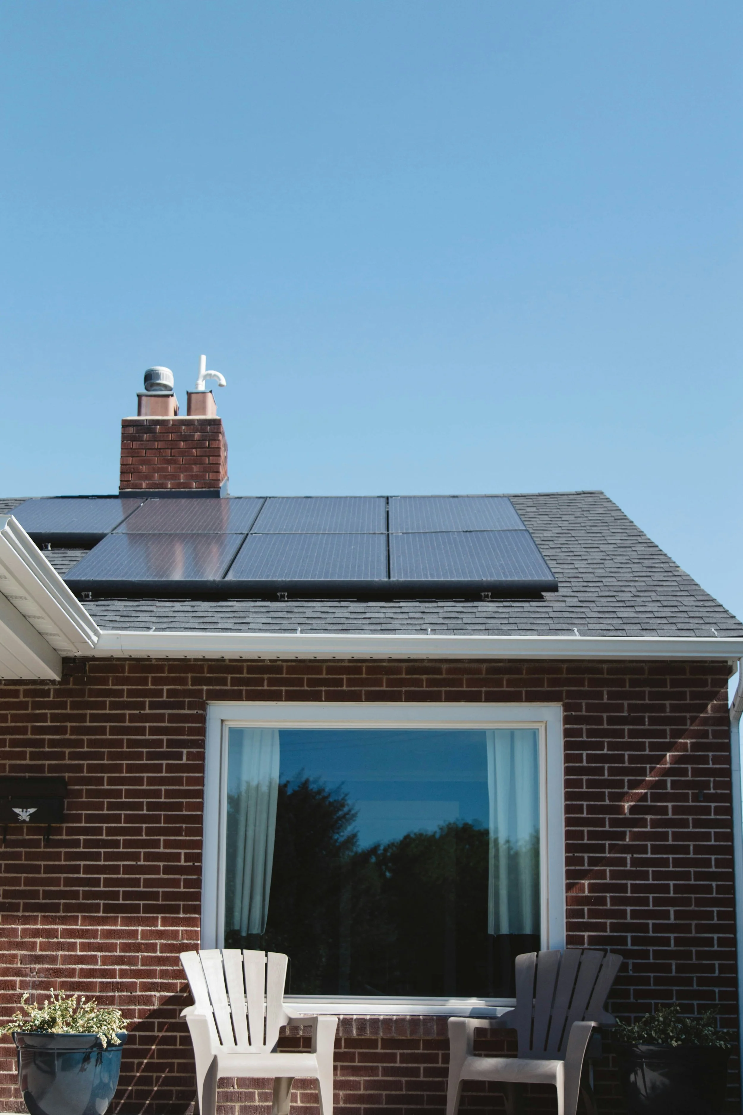 Brick house with solar panels on roof, chimney, large window, two plastic chairs, and plant in pot.