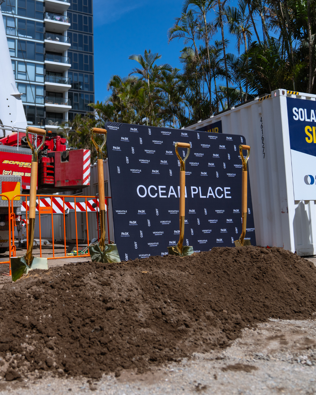 Construction site with a blue Ocean Place sign, orange safety barriers, a pile of dirt, and construction equipment. Tall building and palm trees in the background.