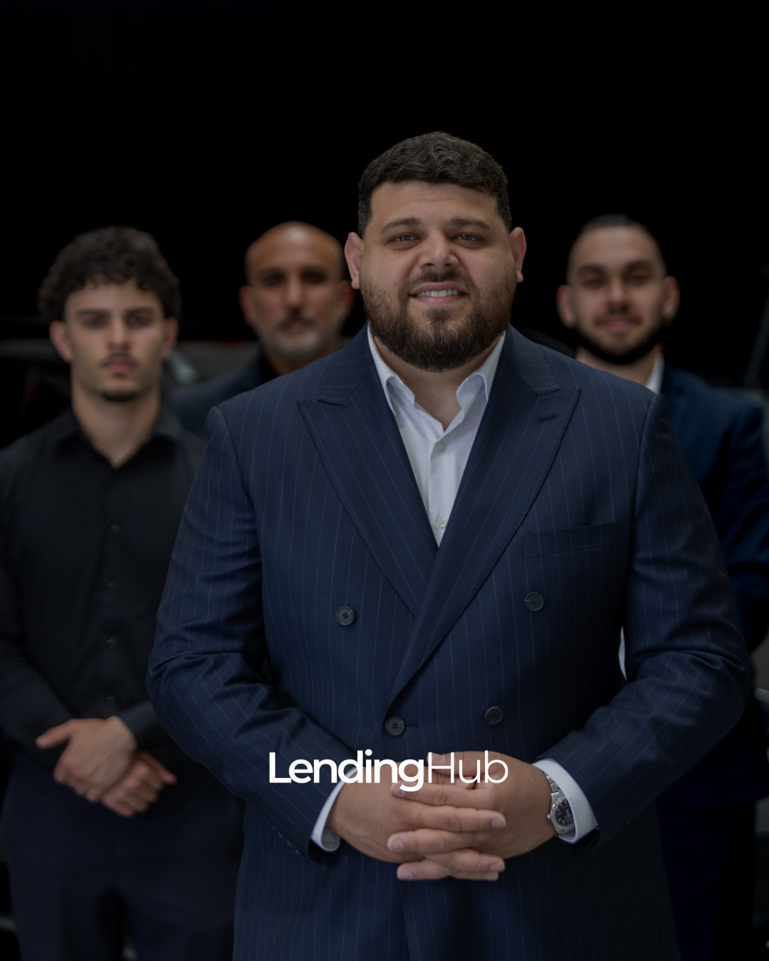 A group of four men in suits standing in a black background, with the man in front smiling, wearing a dark pinstripe suit and white shirt, with the LendingHub logo on the image.