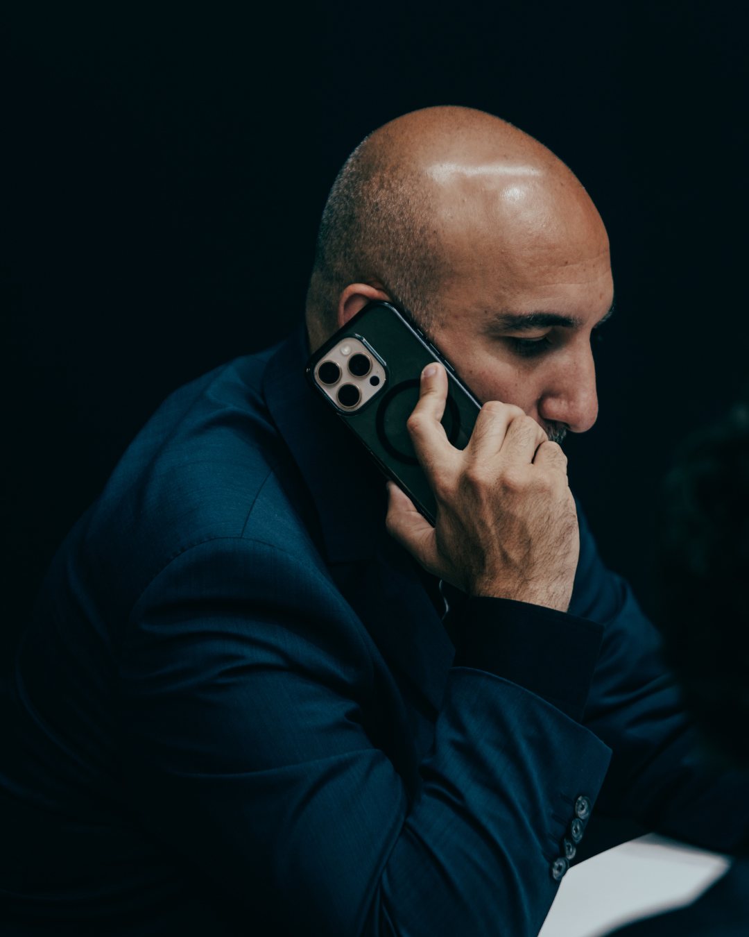 A man with a shaved head wearing a dark blue suit, talking on a smartphone with a black protective case, in a dimly lit environment.
