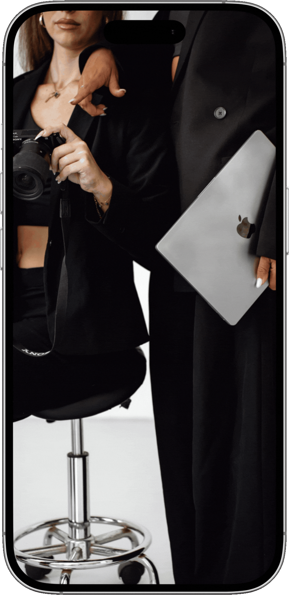 A woman taking a mirror selfie with a camera, holding a laptop in her left hand. She is dressed in black and wearing jewelry.