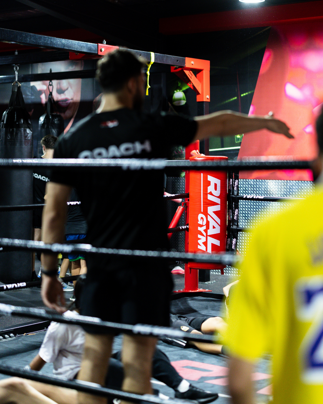 A boxing gym with people training inside, including a trainer in black holding a stretching position in a boxing ring.