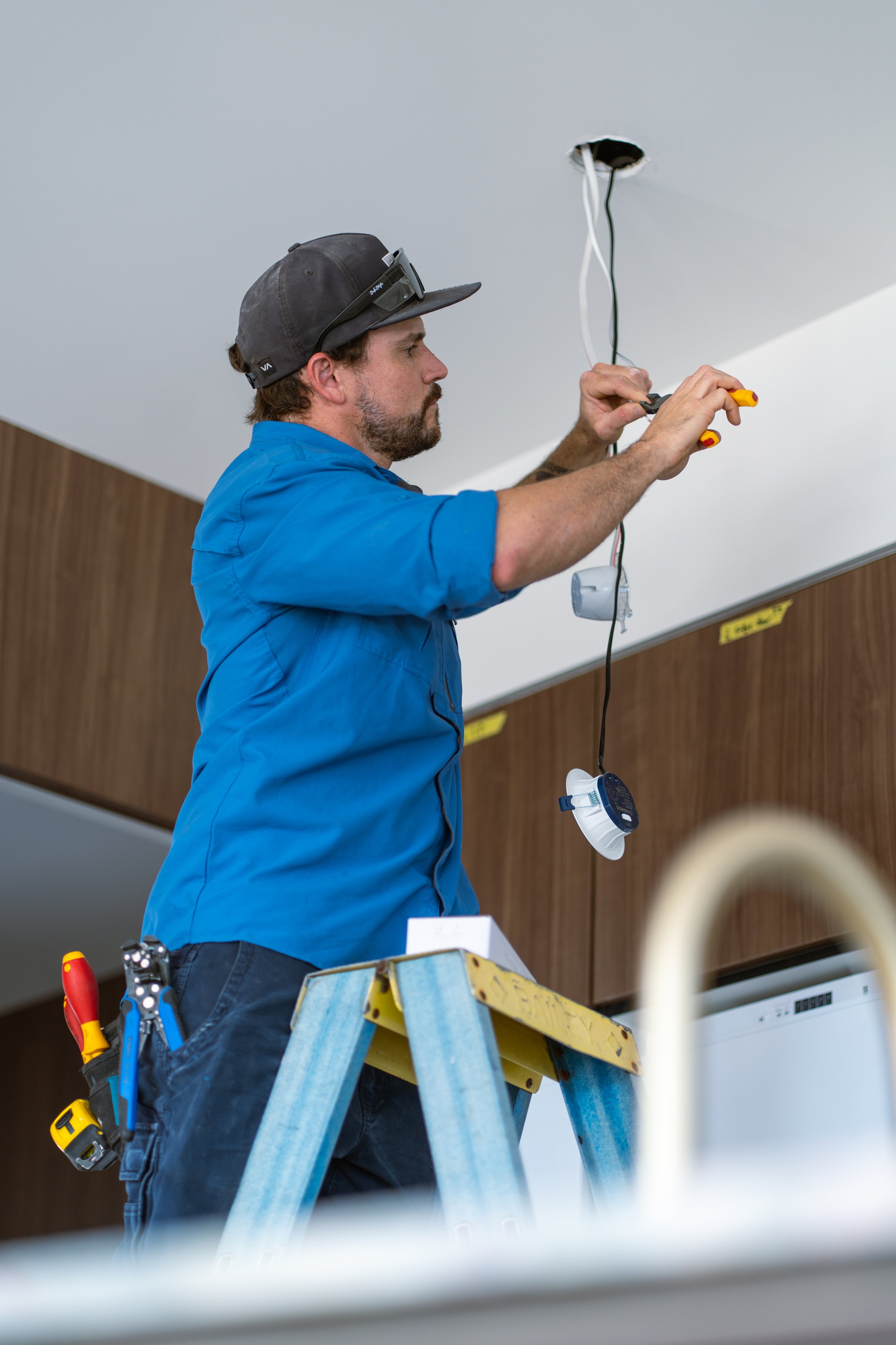 A man working on a ceiling, using tools and electrical wires, standing on a ladder.