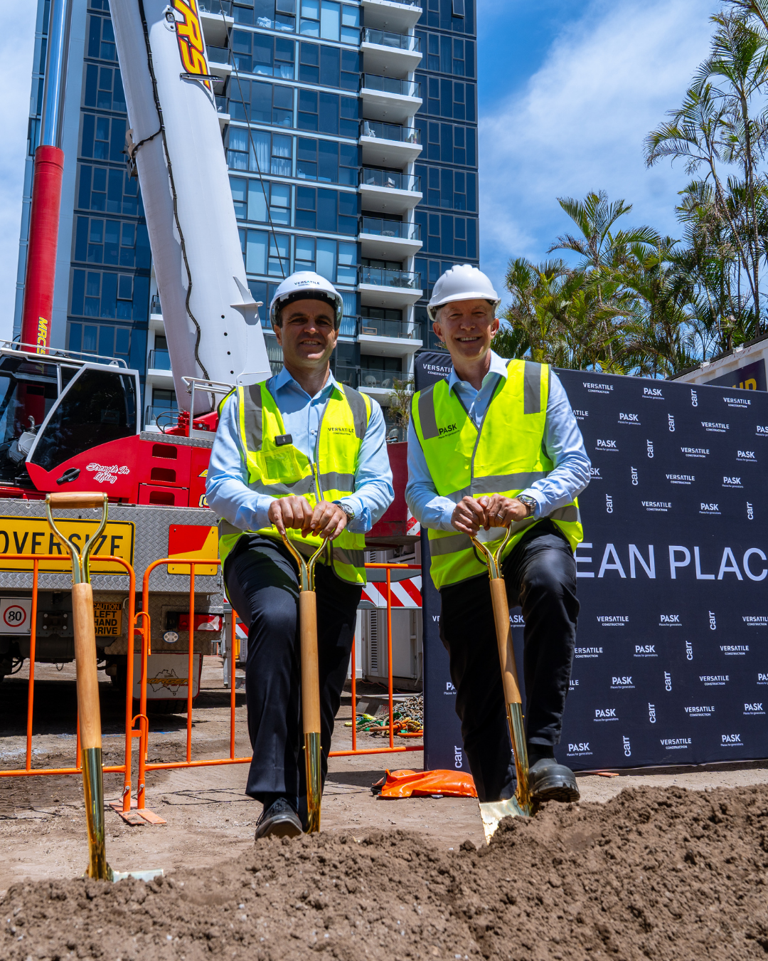 Two men in safety vests and hard hats at a construction site, holding shovels, with a high-rise building, construction equipment, and palm trees in the background.