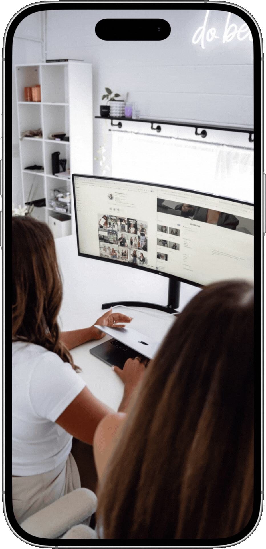 Two women sitting at a desk with a large curved computer monitor, working together on social media or a digital project, in a bright, modern room with white decor and bookshelf.