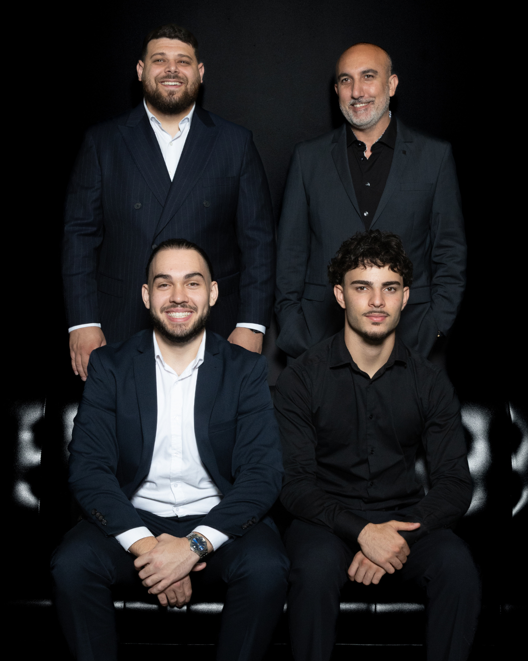 Four men in formal suits posing for a photo against a black background, with two sitting on a black leather sofa and two standing behind them.