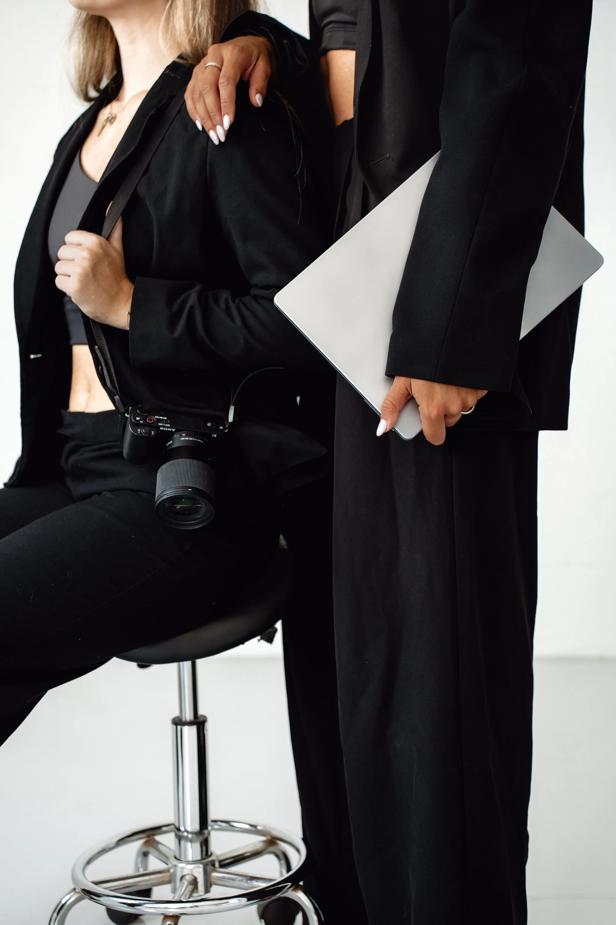 Two people dressed in black, one seated with a camera hanging around their neck and the other standing holding a closed laptop.