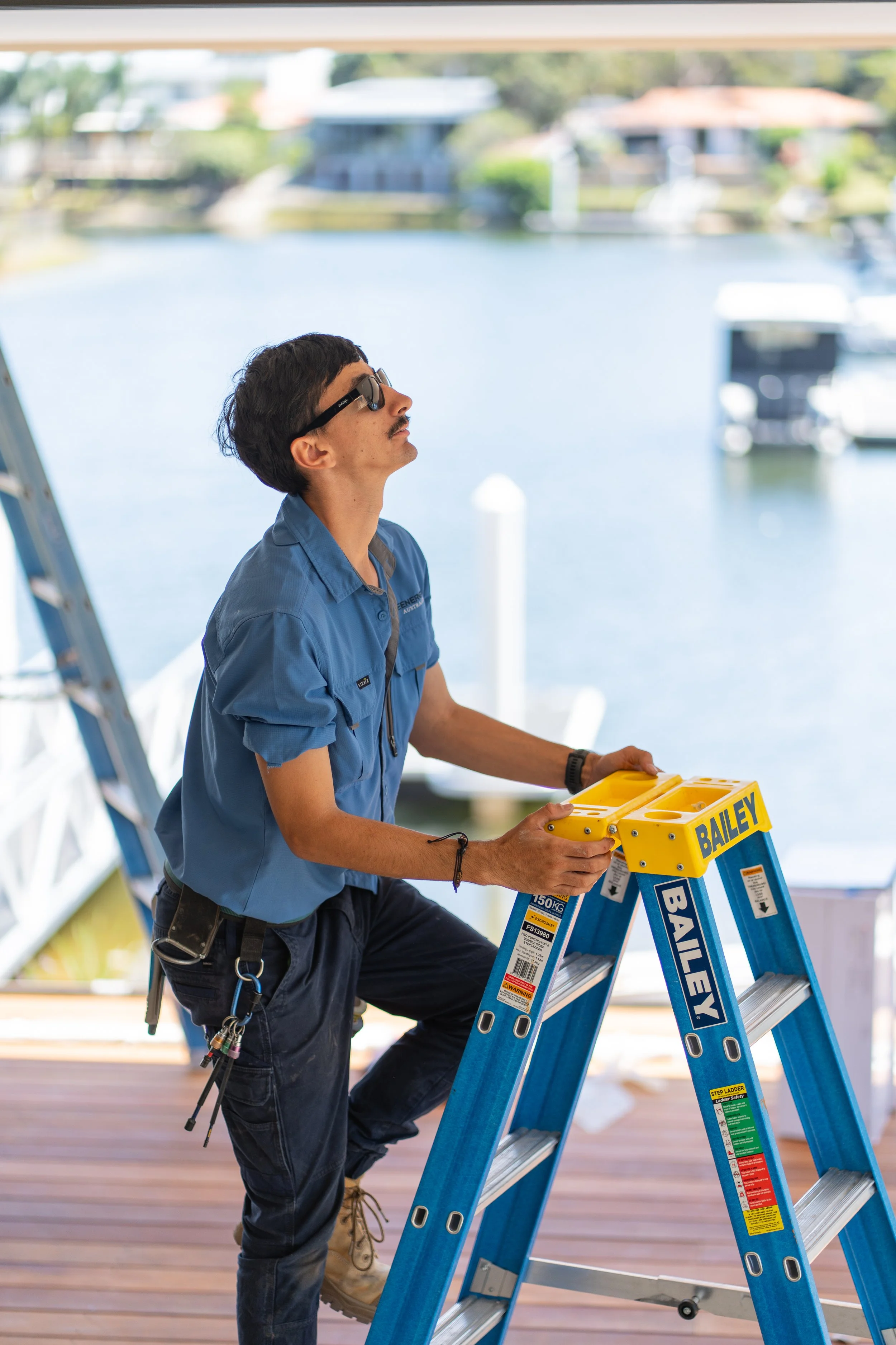 A man standing on a wooden deck near water, holding a yellow Spirit level, wearing sunglasses and a blue work shirt.