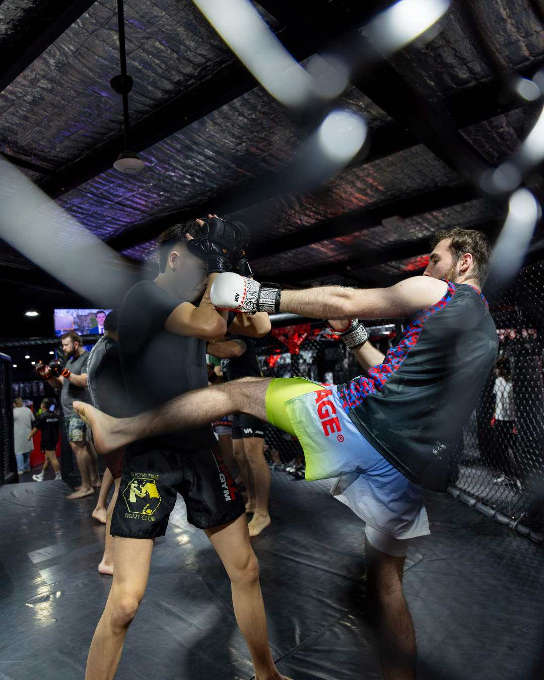 Two men practicing mixed martial arts training inside a cage. One man is executing a kick while the other is blocking with his hands up.