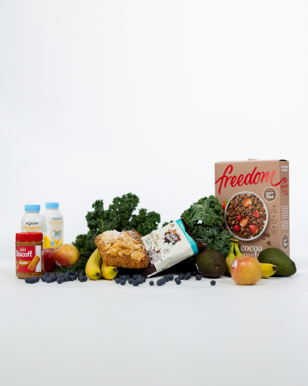 Collection of healthy foods including blueberries, kale, bananas, apples, avocados, a muffin, protein supplements, and a box of cocoa crunch cereal on a white surface against a white background.